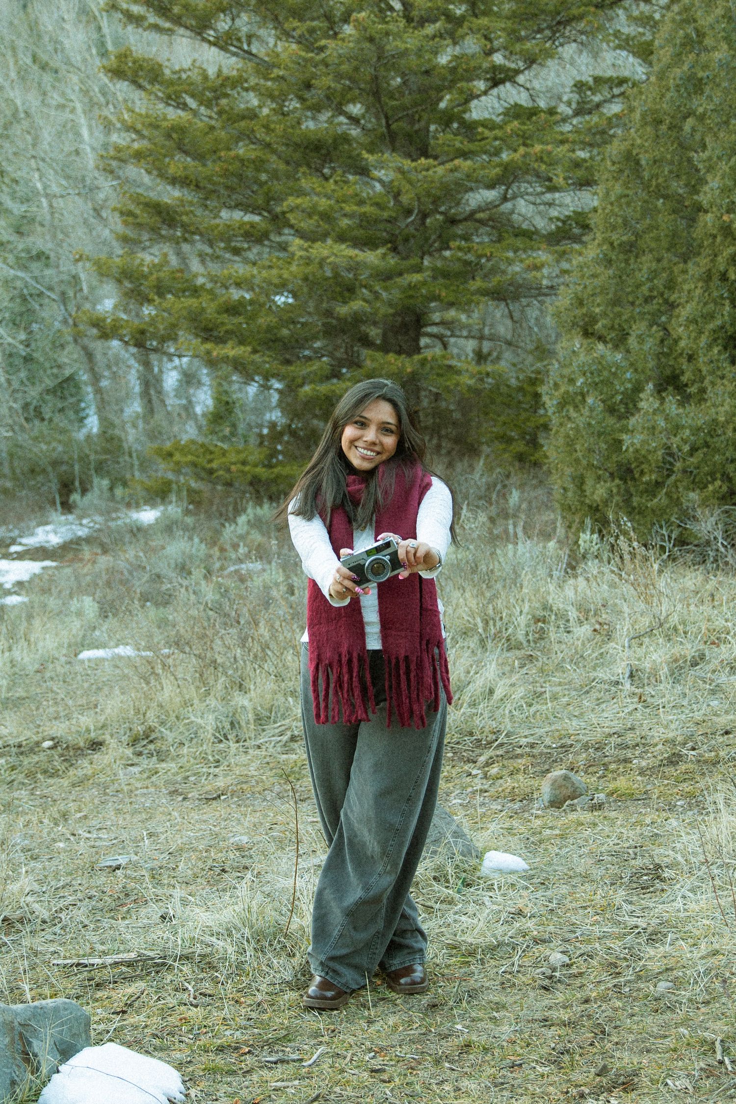 A person in a burgundy vest and gray pants smiles while standing on a snowy hillside among evergreen trees.