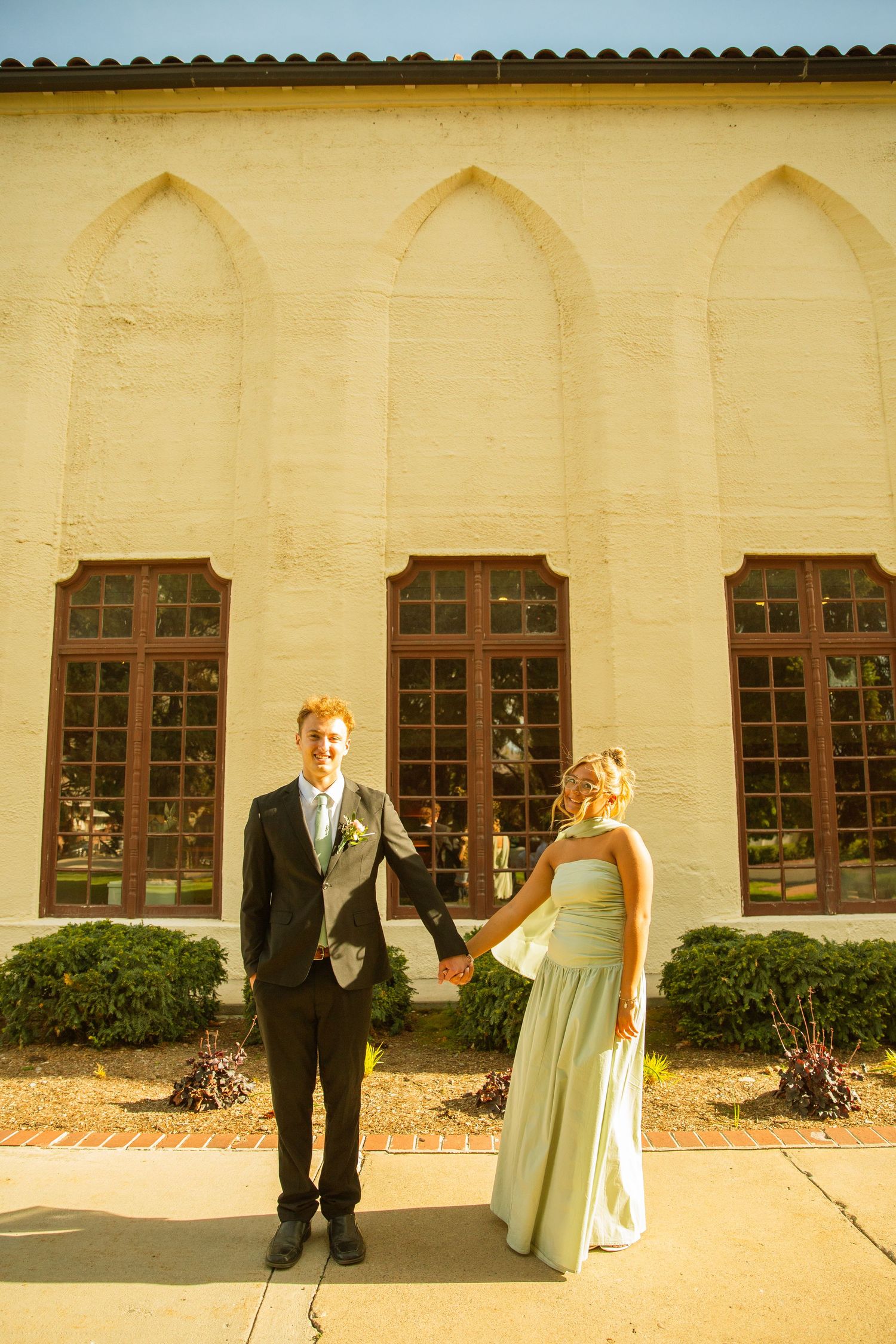 Young couple in formal attire holding hands outside a historic cream-colored building with arched windows on a sunny day.