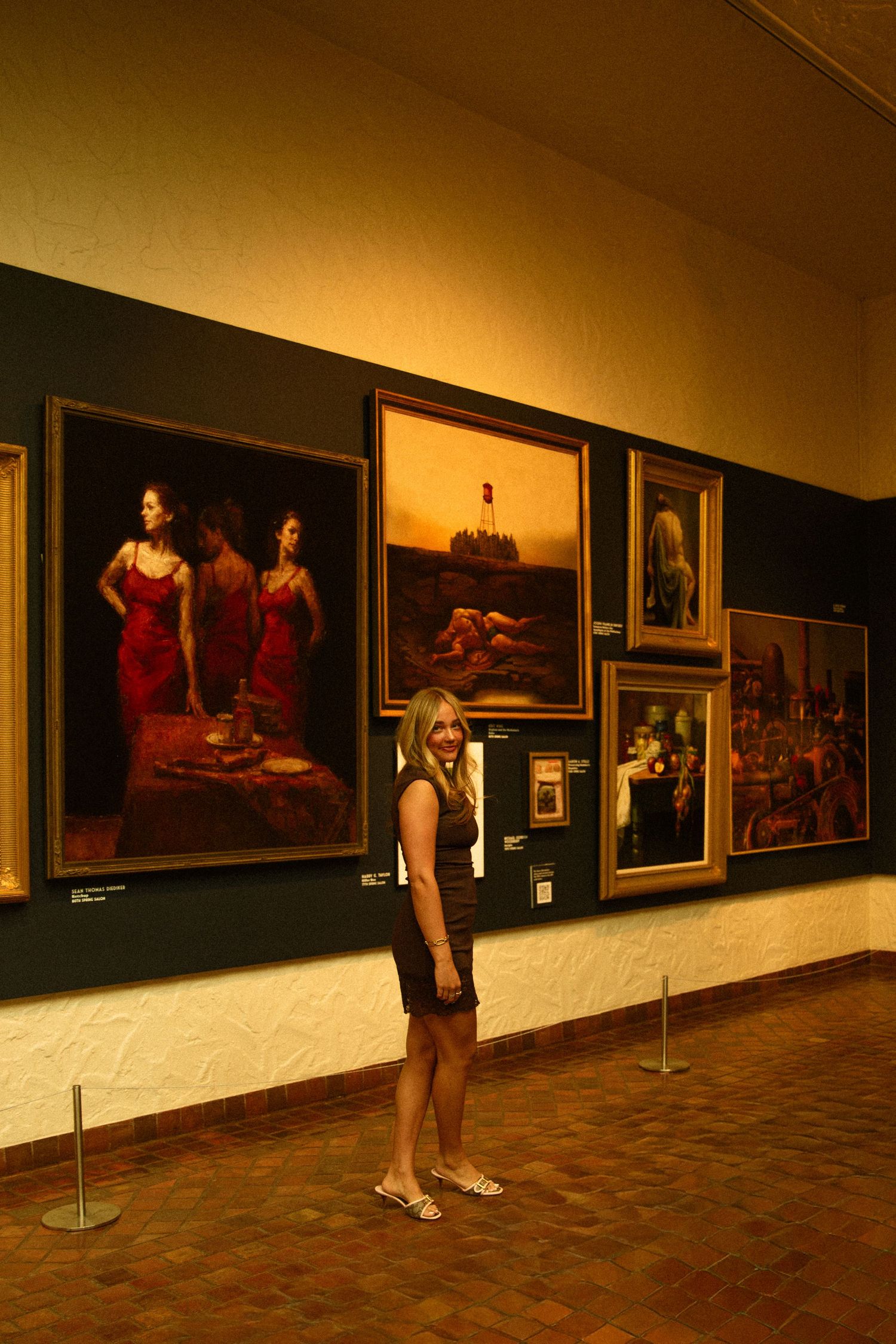 Blonde woman in black dress standing in art gallery beside large oil paintings of women in red dresses.