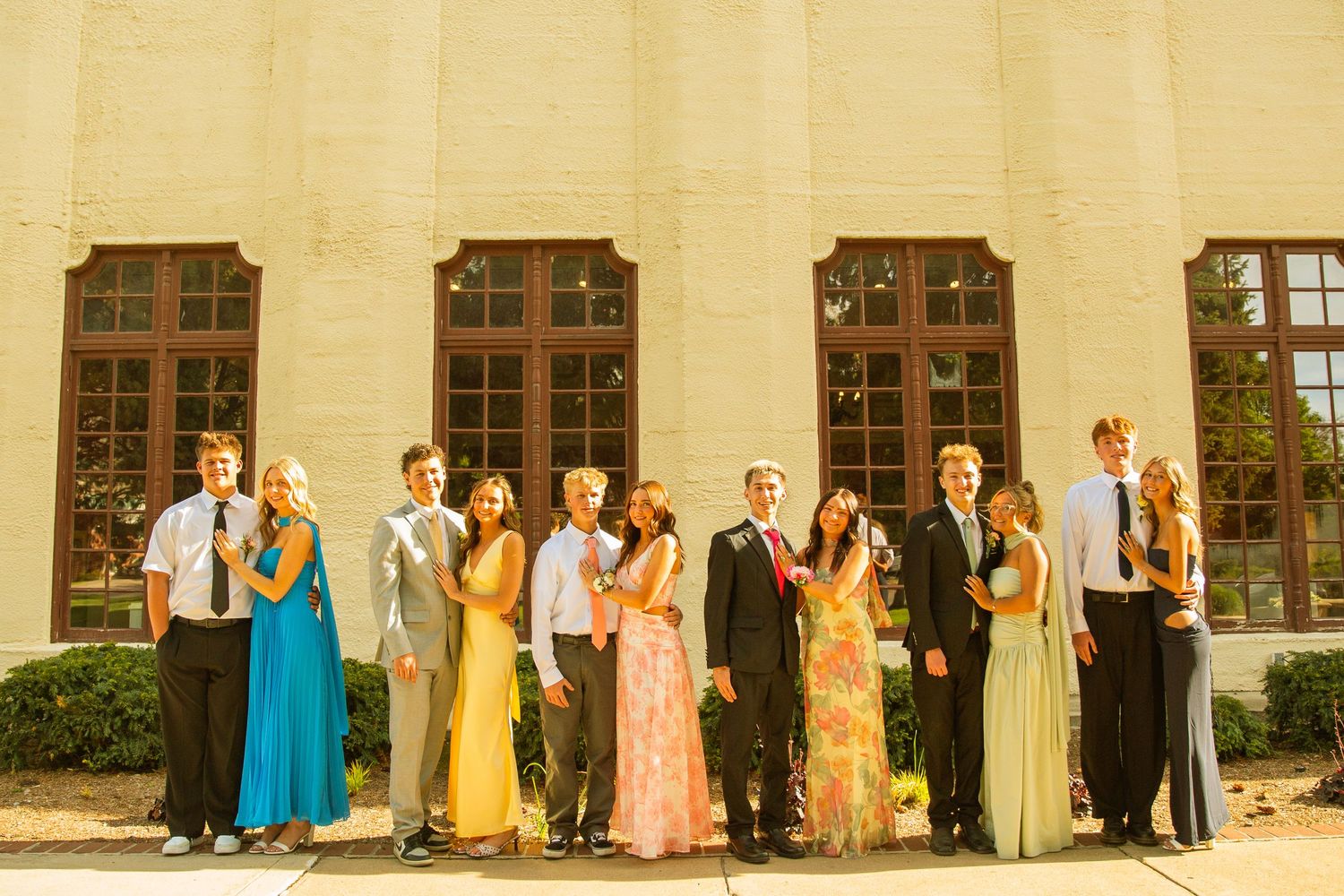 Group of teens in formal prom attire posing outdoors in front of a cream brick building with large windows.
