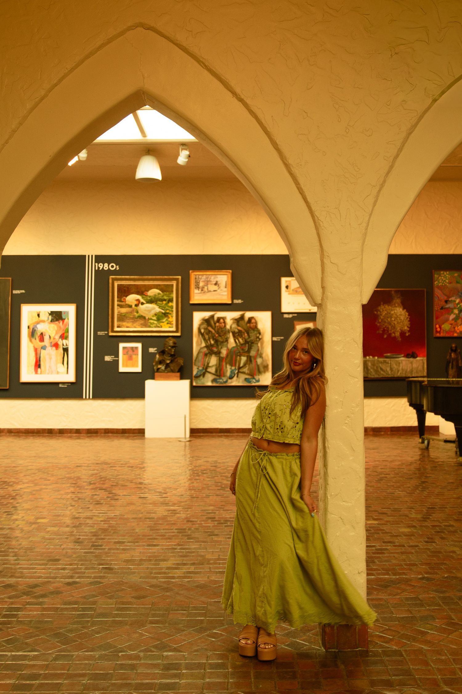Woman in green maxi skirt poses in art gallery with arched ceilings and colorful paintings on display.