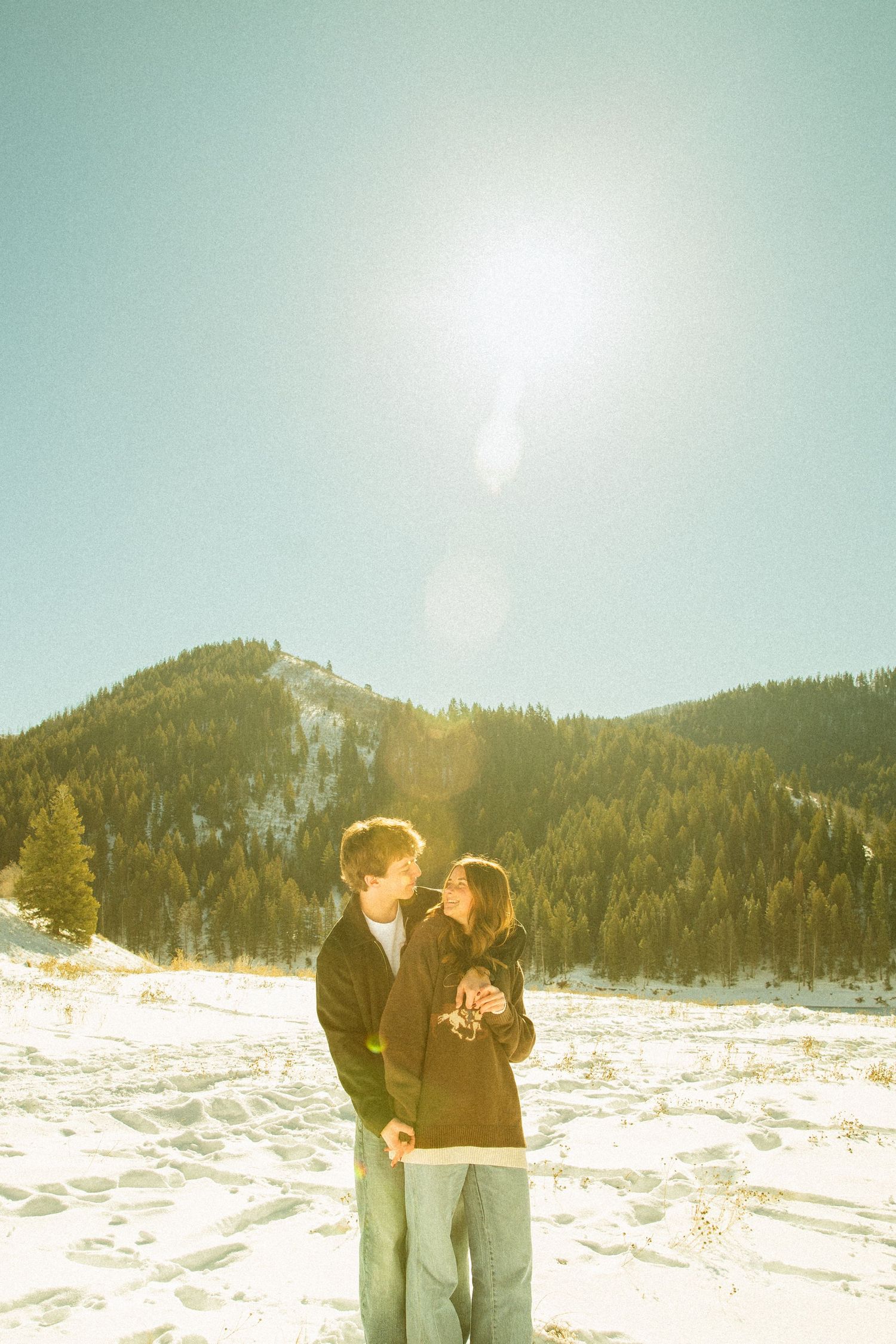 A couple shares a romantic moment in a snowy mountain landscape with bright sunlight streaming through the winter scene.