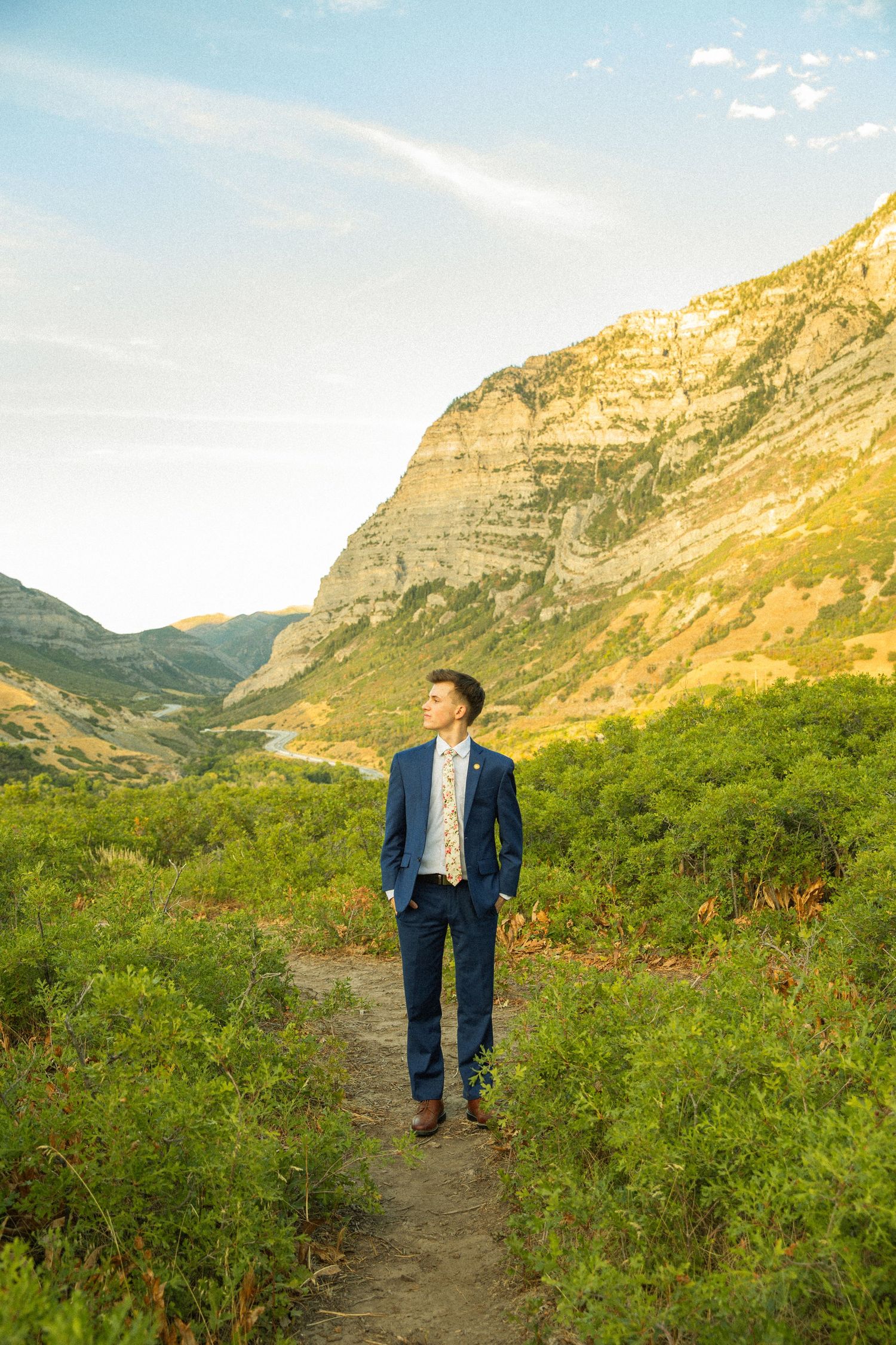 Someone in a navy suit walks along a mountain trail surrounded by rugged cliffs and green vegetation at sunset.