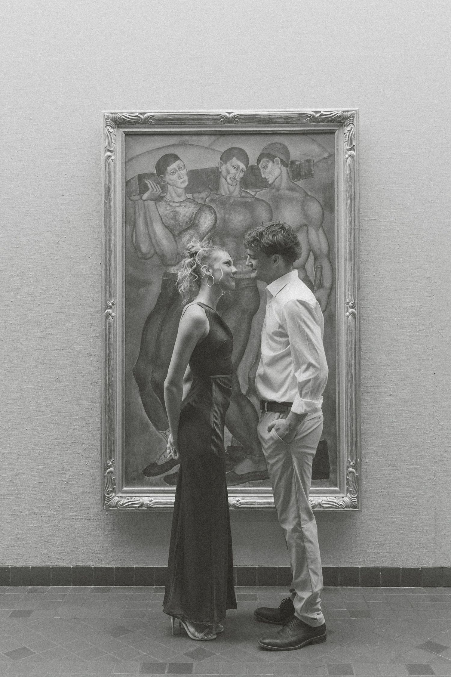 Two people observe and discuss artwork displayed in frames on a gallery wall in this vintage black and white photograph.