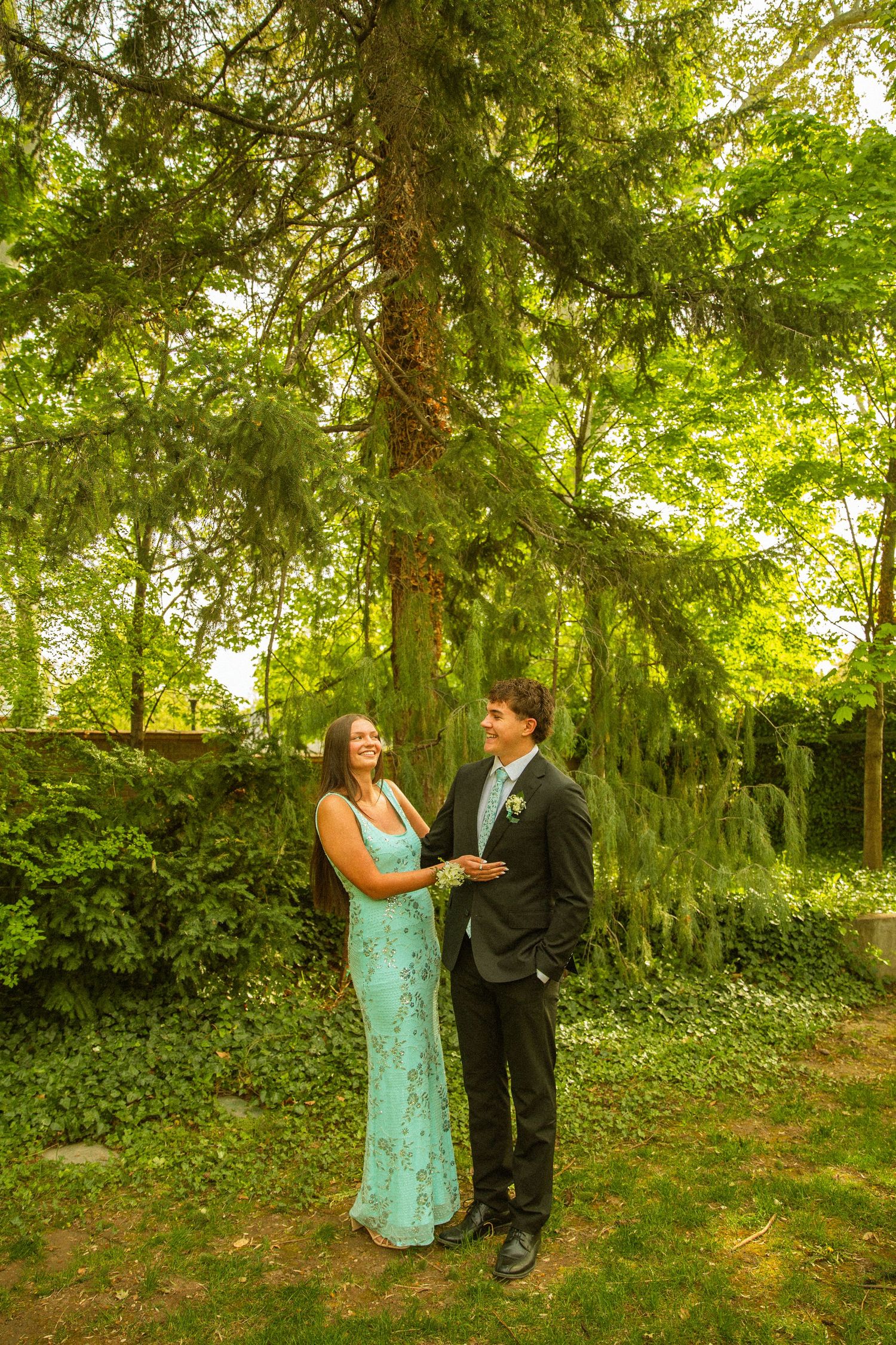 A couple in formal attire stands smiling in a lush green garden, woman in teal gown, man in dark suit with boutonniere.