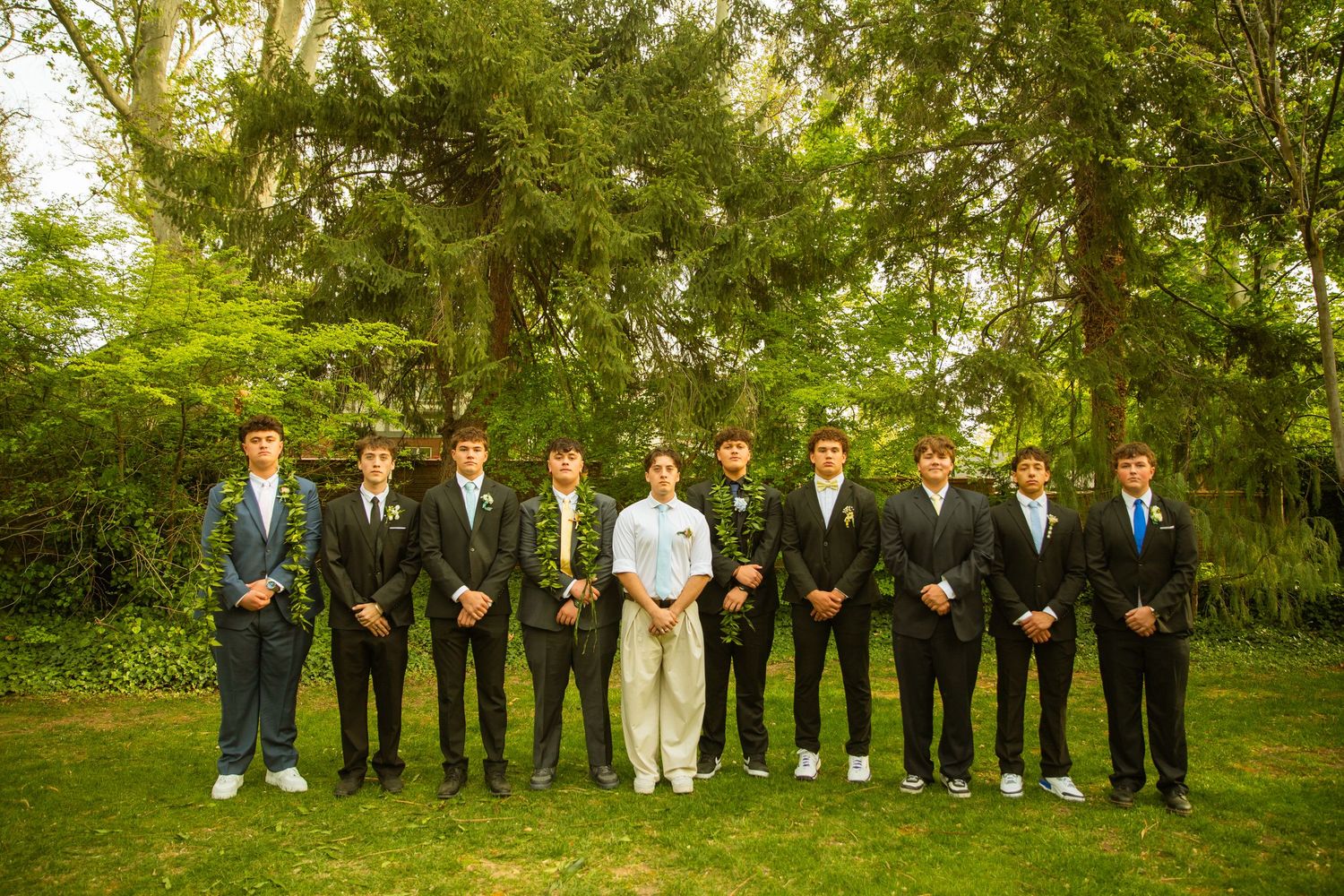Group of young men in formal suits posing outdoors on green grass with lush trees in the background.