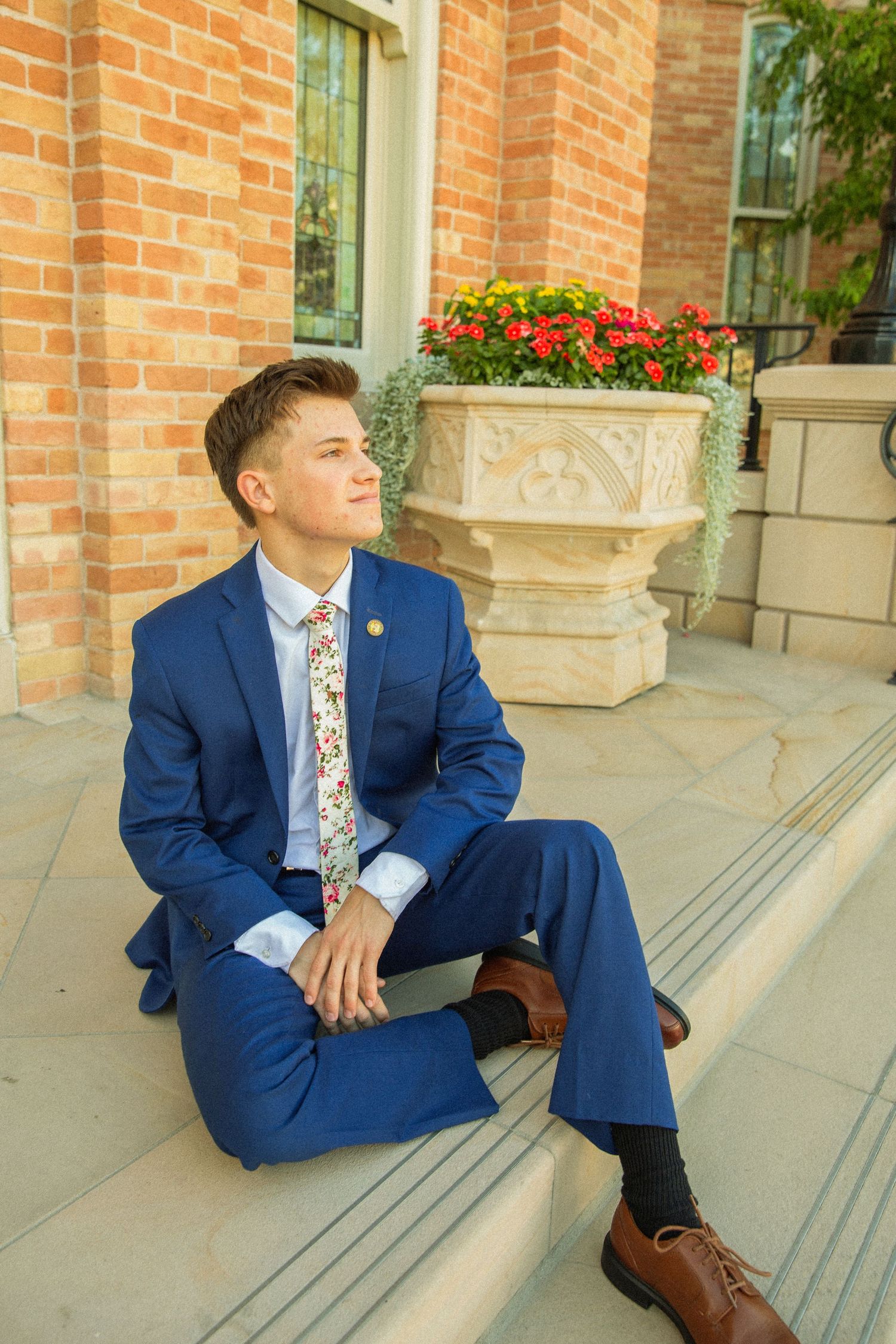 Person in navy blue suit sits on concrete steps outside brick building with floral window boxes.