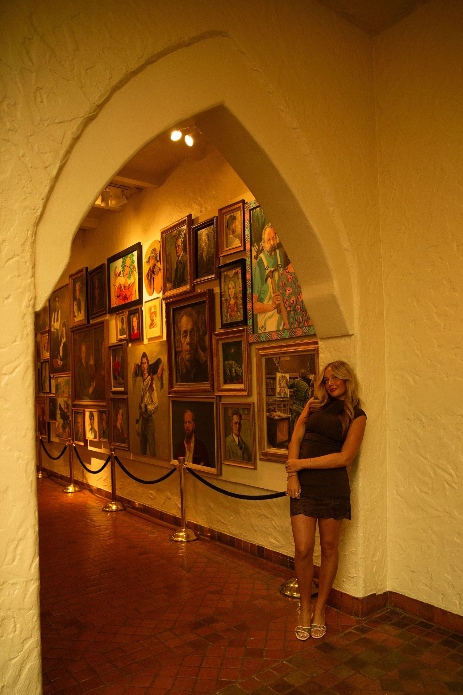Woman in black dress stands beside wall of colorful portrait paintings in arched gallery hallway.