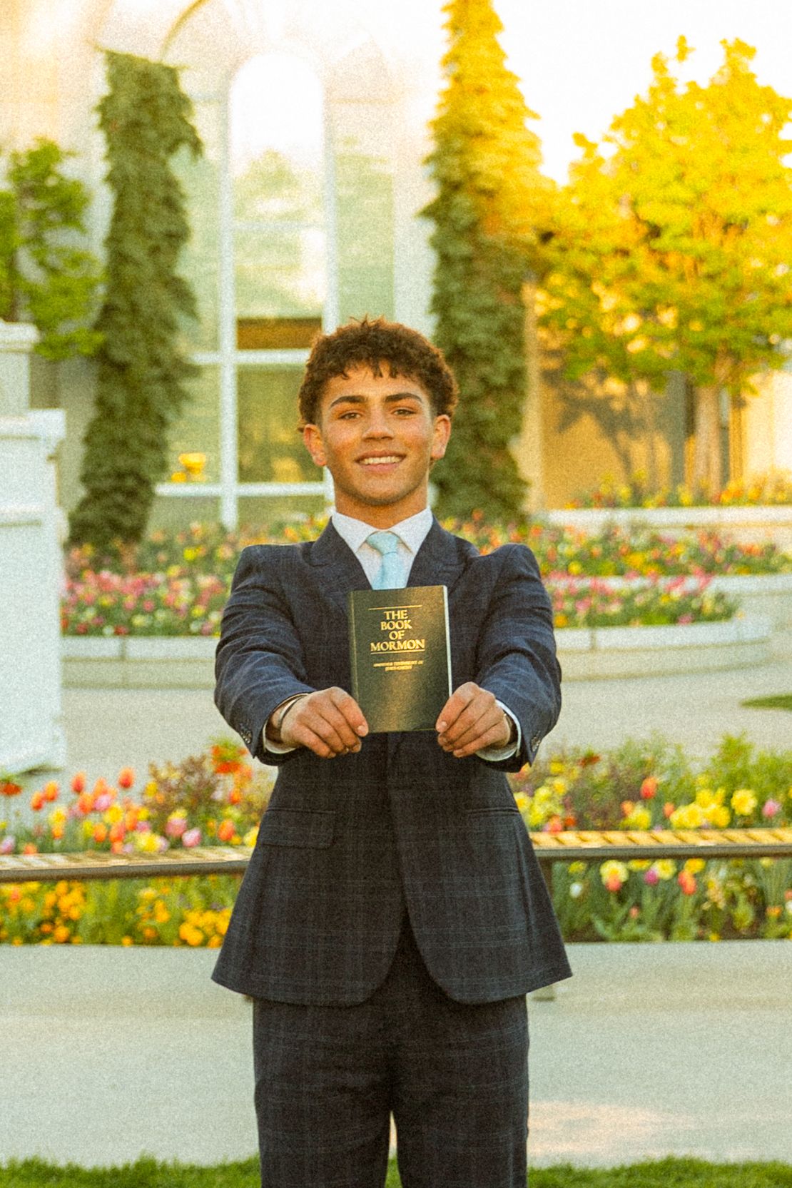 Young man in navy suit smiling while holding Book of Mormon outdoors near colorful garden and trees.