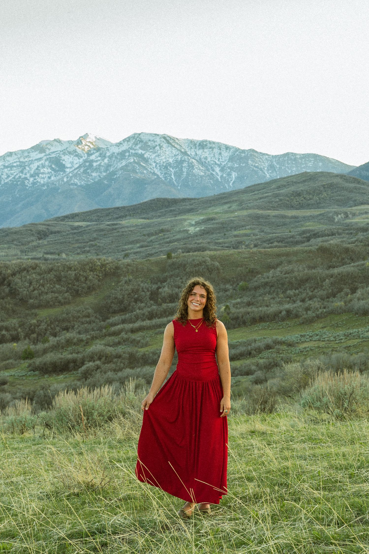 Woman in red maxi dress standing in green mountain meadow with snow-capped peaks in background.