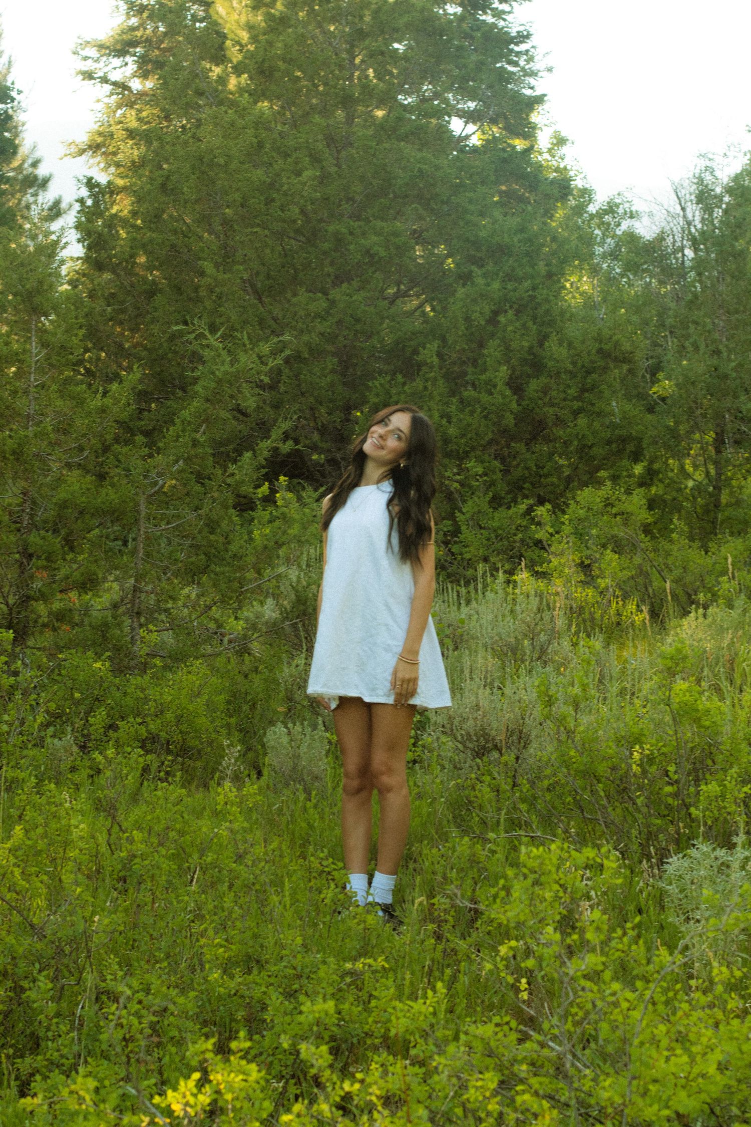 Person in white clothing standing among wildflowers in a sunny forest clearing.
