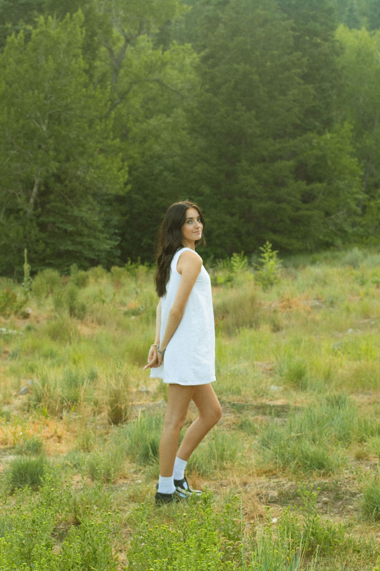 A person in a white sleeveless top walks through a grassy meadow with evergreen trees in the background.