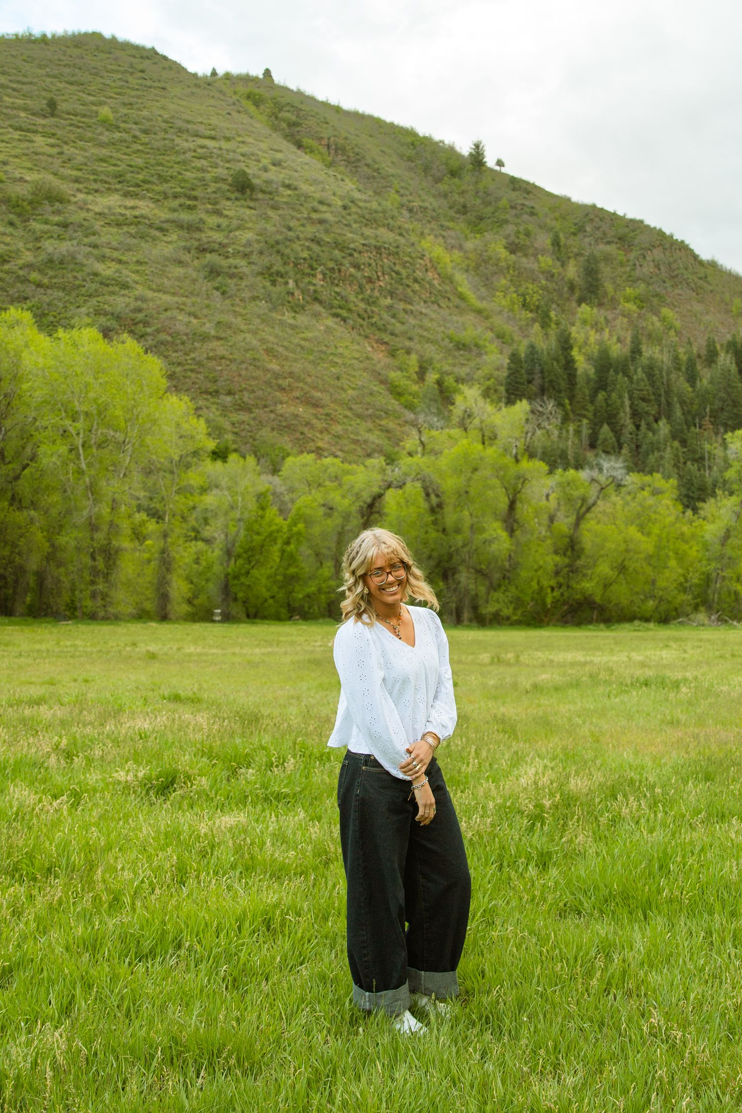 Woman in white blouse and black pants smiling in green meadow with forested mountain backdrop.
