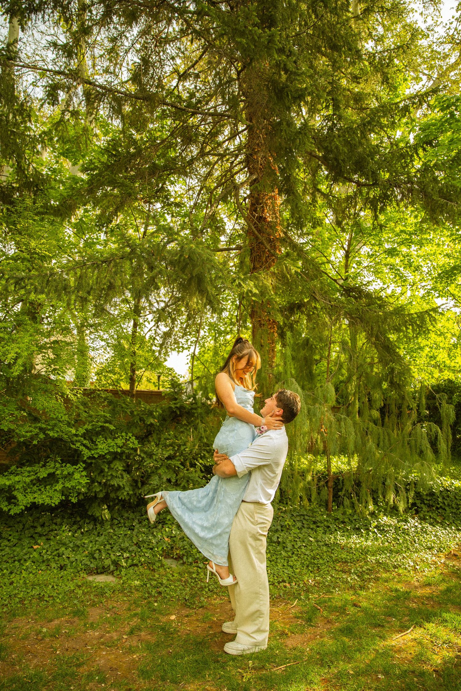 A man lifts a woman in a blue dress outdoors under a large green tree in a romantic garden setting.