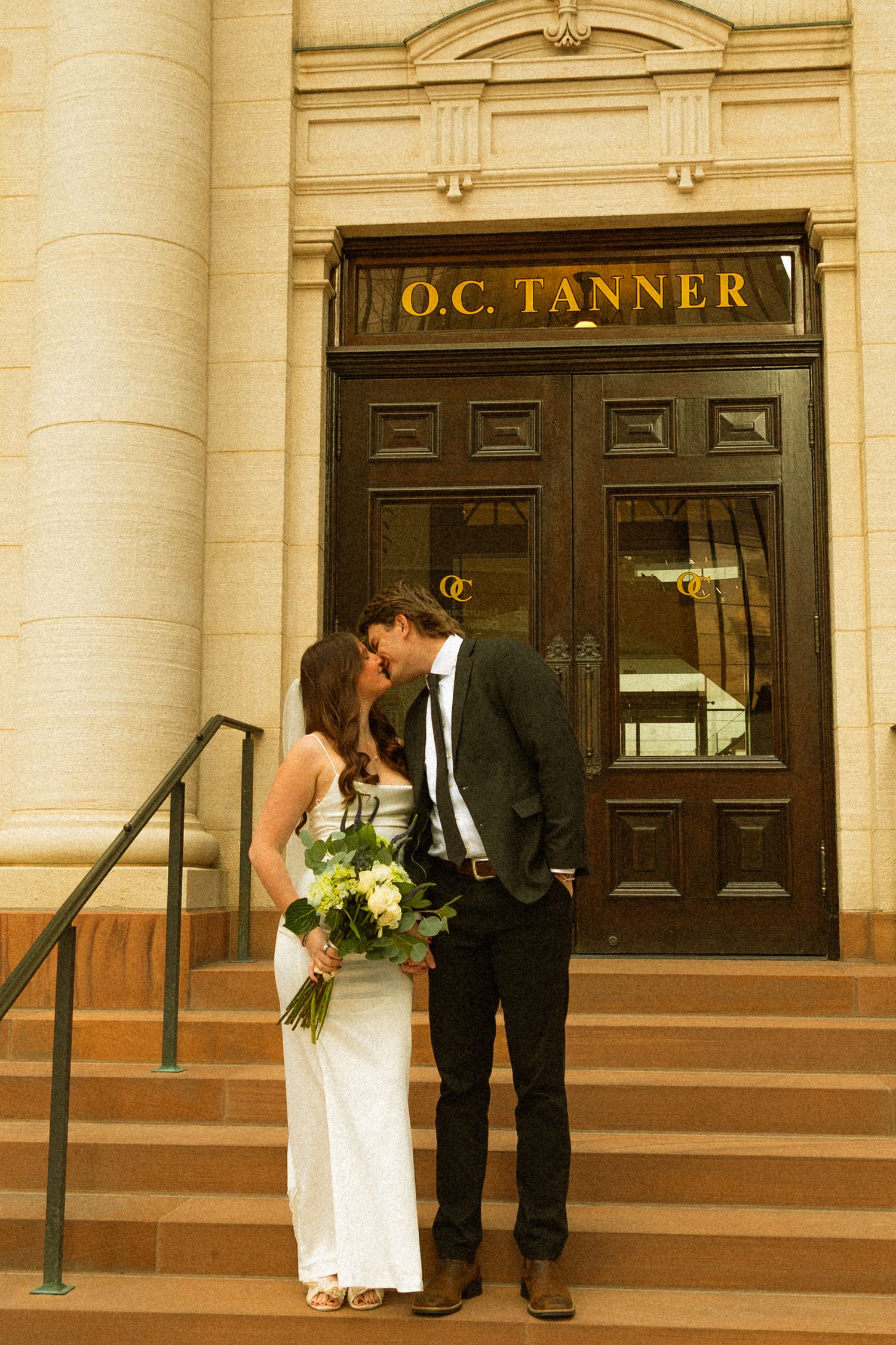 A couple shares a romantic moment on the steps of the O.C. Tanner building, with the bride holding a yellow bouquet.