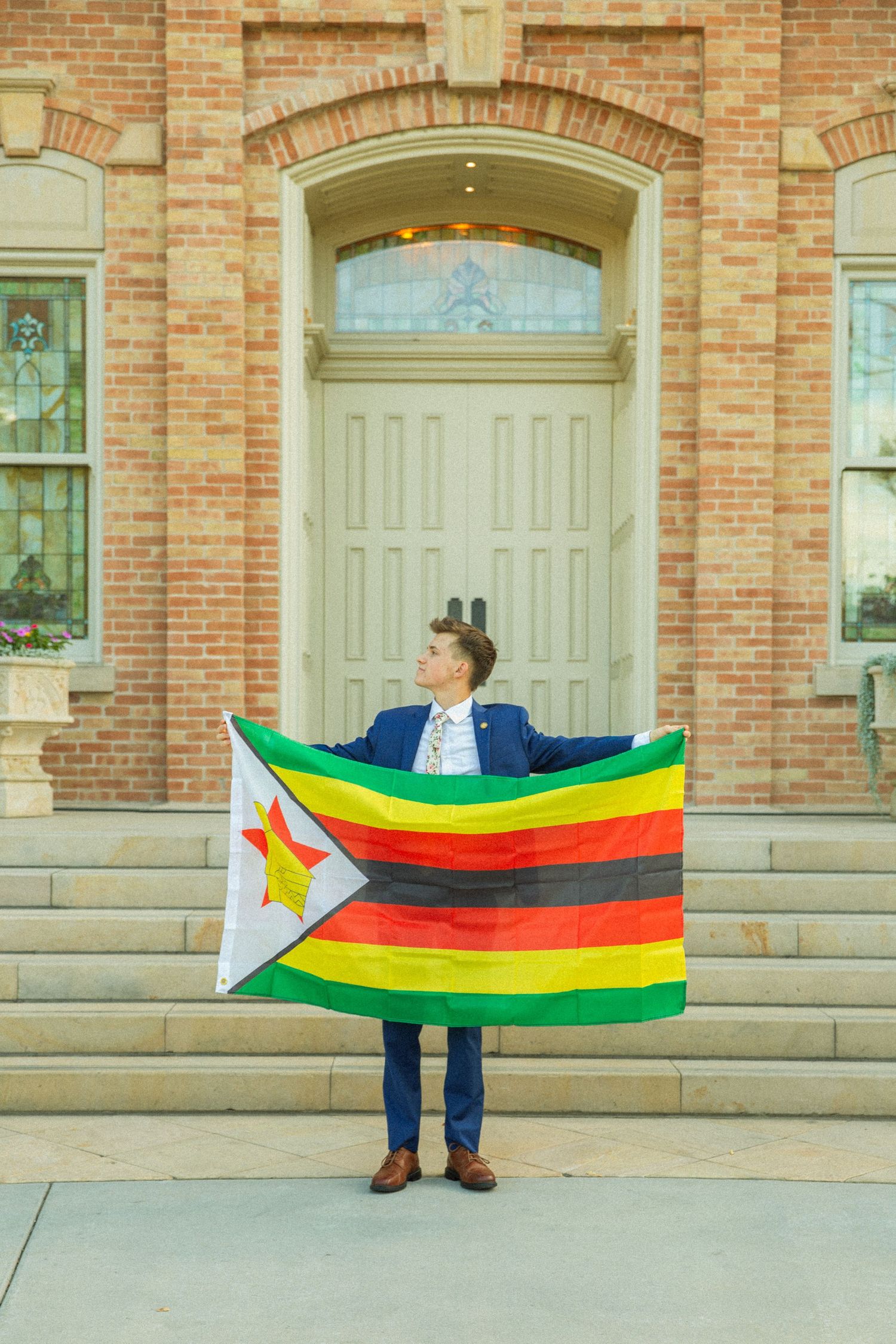 Someone in formal attire holds up the Zimbabwe national flag in front of a historic brick building with arched doorway.