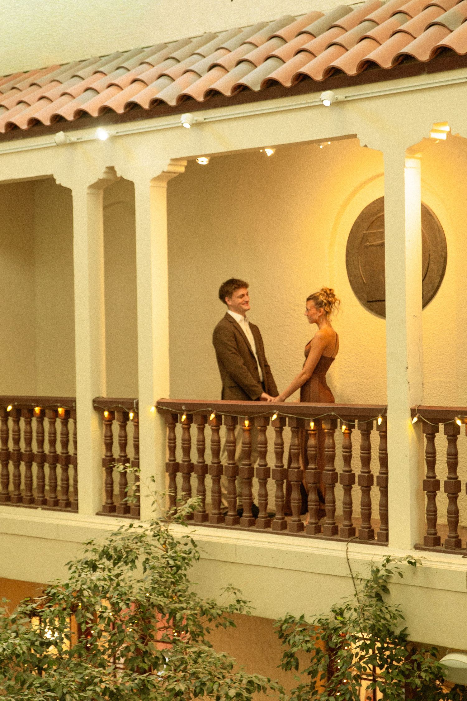 A couple stands together on a Spanish-style balcony with terracotta roof tiles and white columns at dusk.