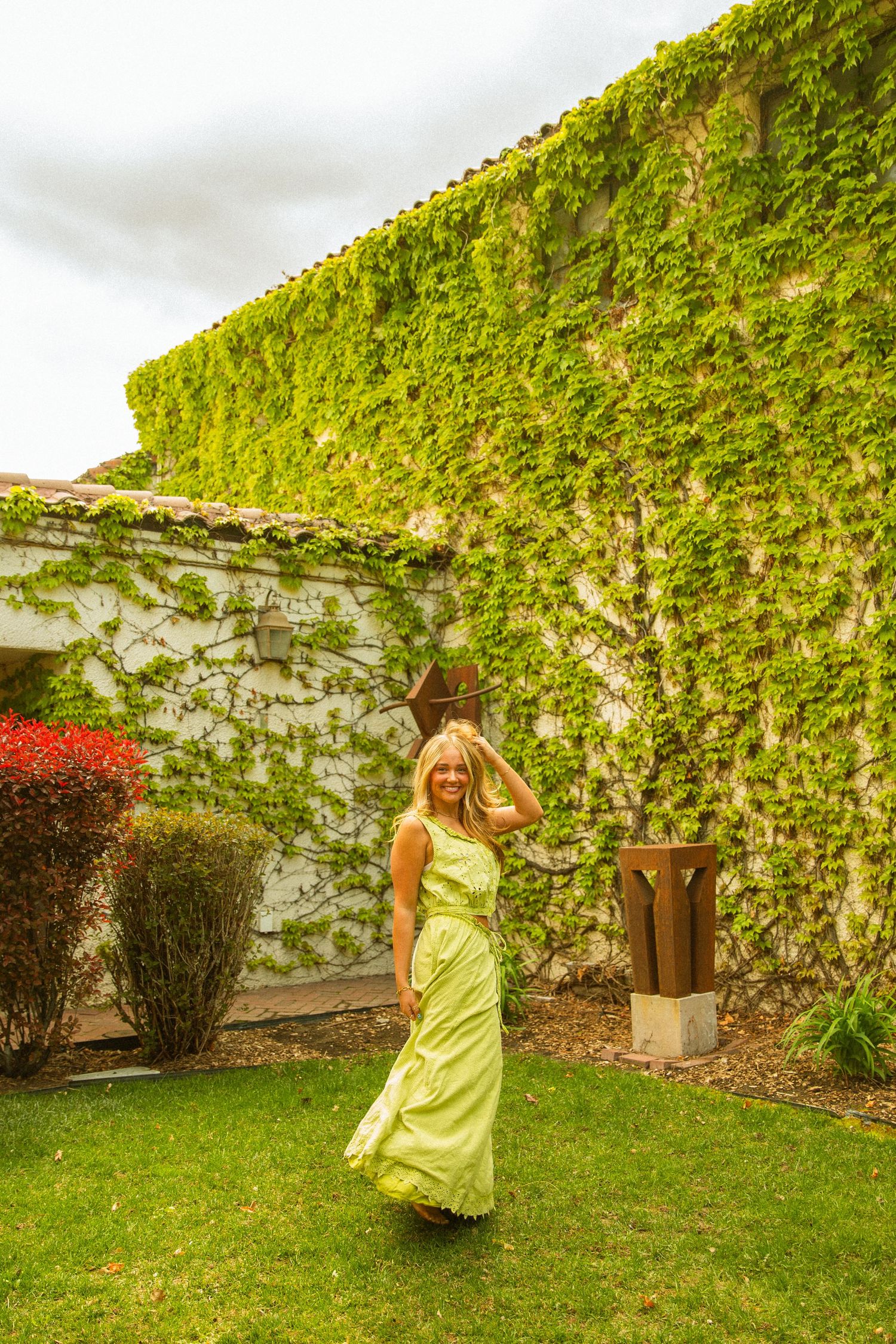 Woman in flowing yellow-green maxi dress poses in lush garden courtyard with ivy-covered stone walls.