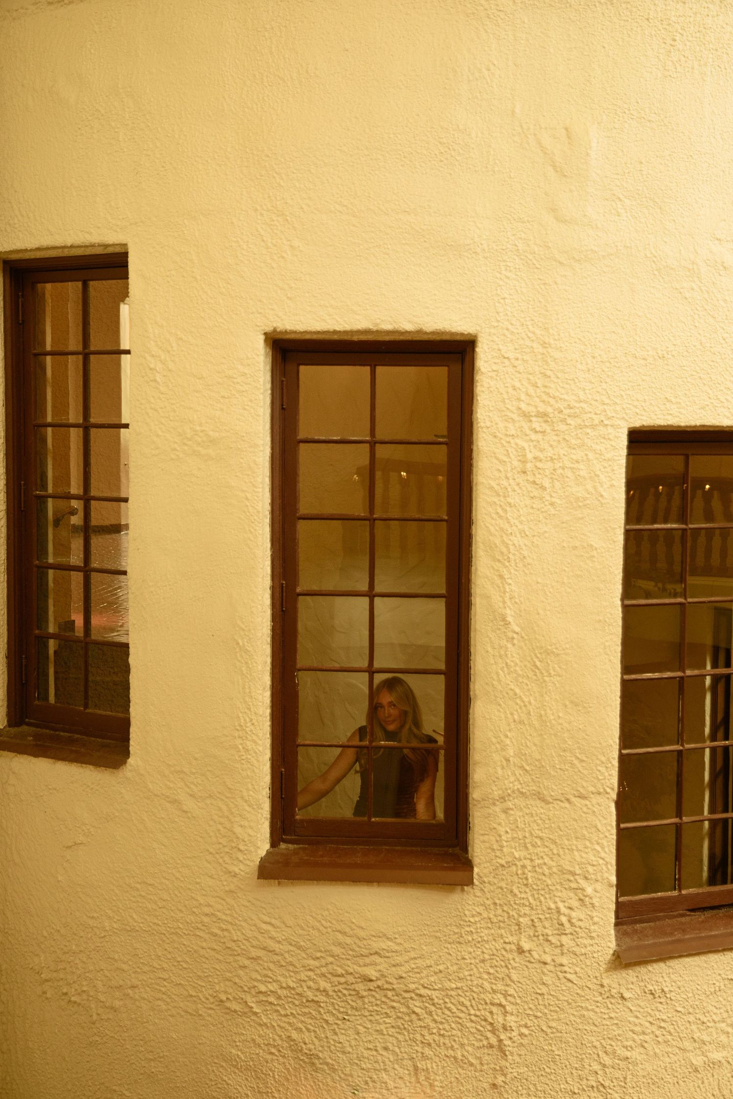 Young woman looking through a wooden-framed window in a textured cream-colored building wall.