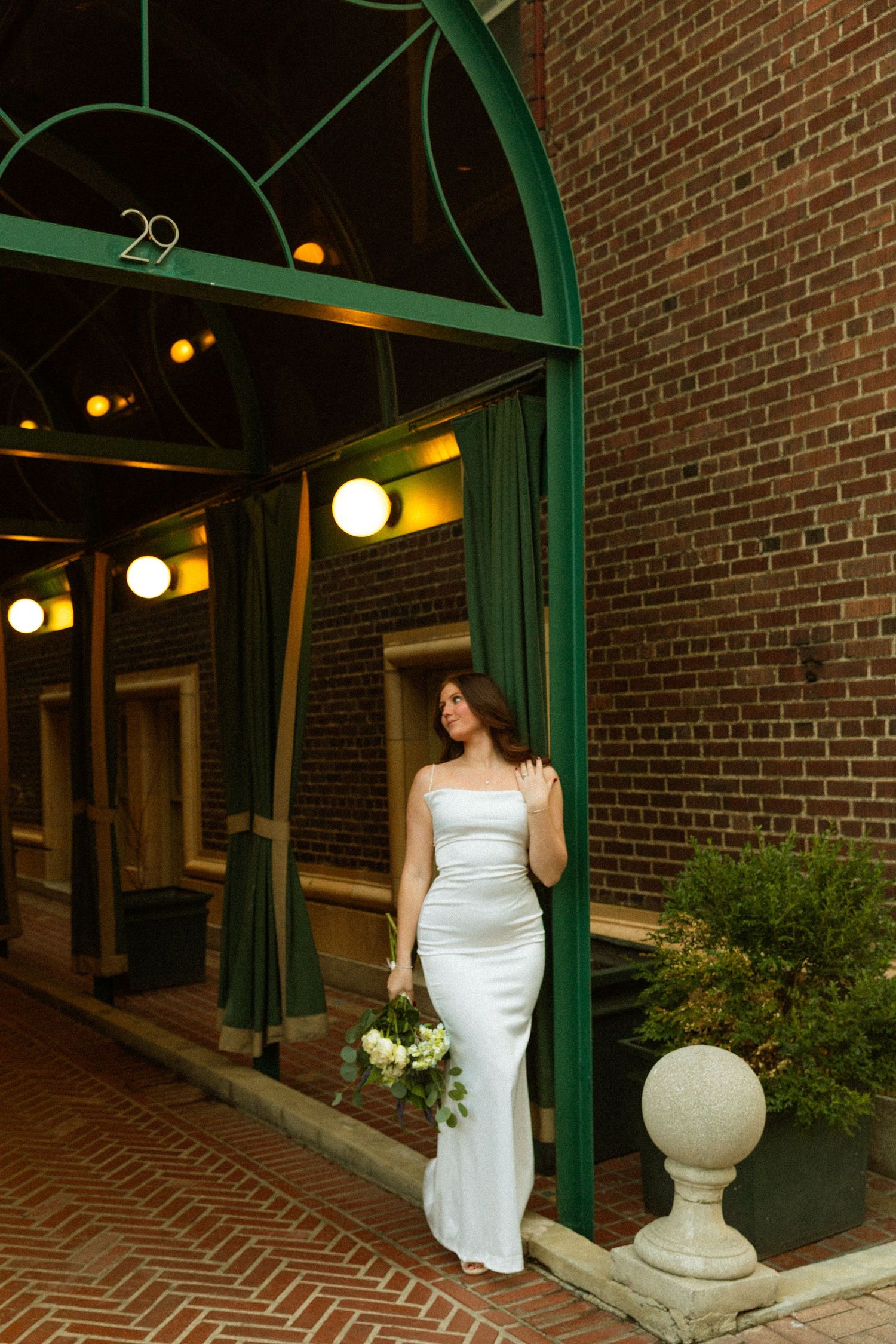 A bride in a sleek white wedding gown stands under a green arched entrance against a brick wall with glowing lights.