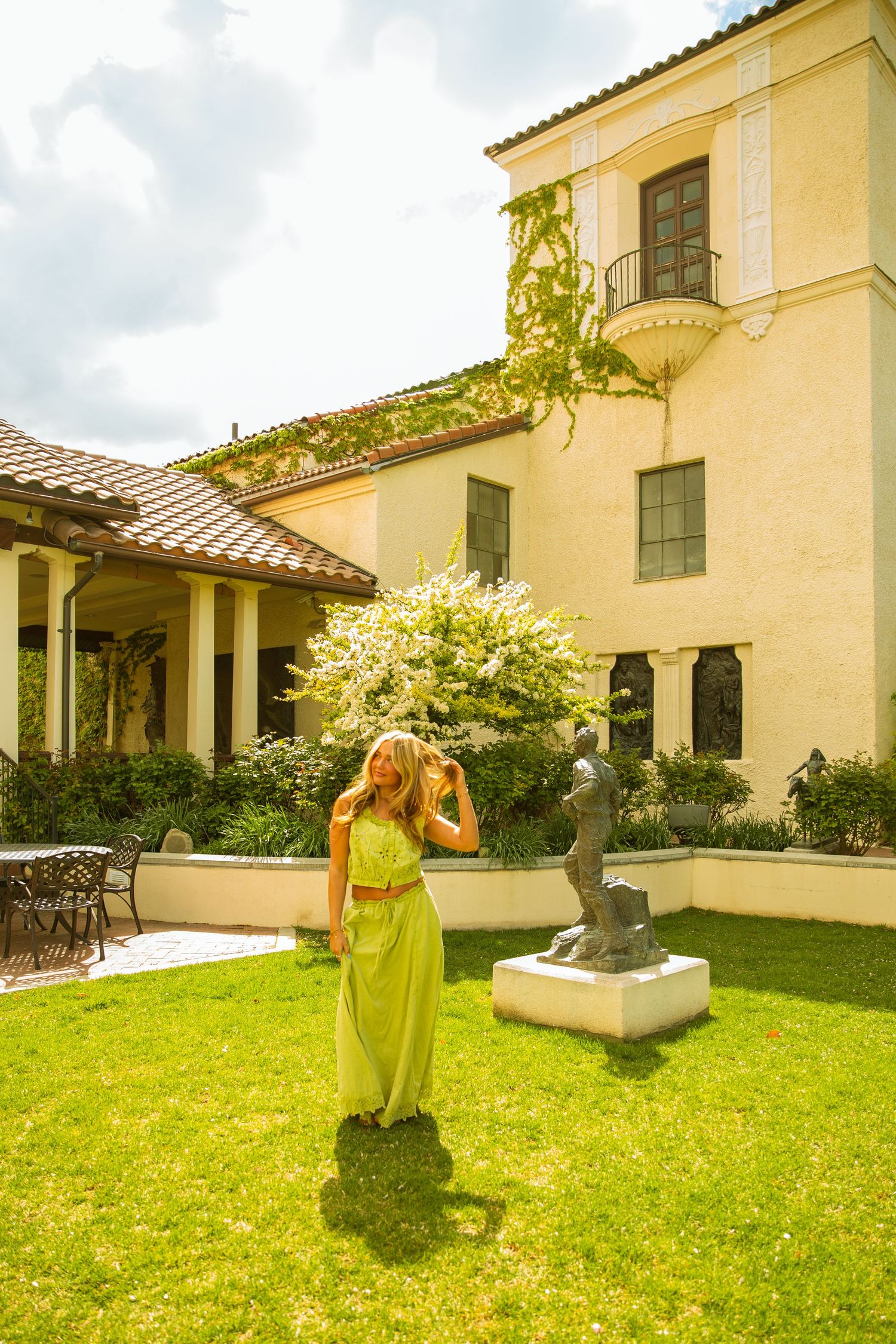 Woman in green maxi dress poses in lush garden courtyard beside bronze sculpture near elegant Mediterranean-style mansion.