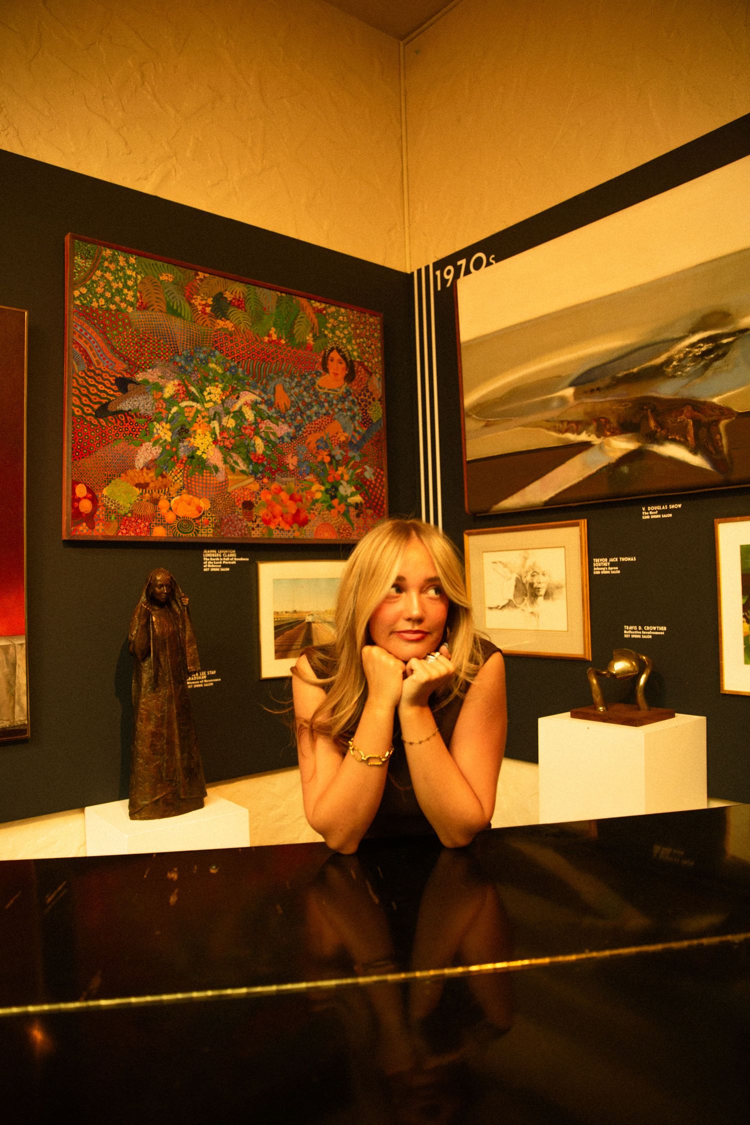 Blonde woman posing in art gallery surrounded by colorful paintings and sculptures in warm lighting.