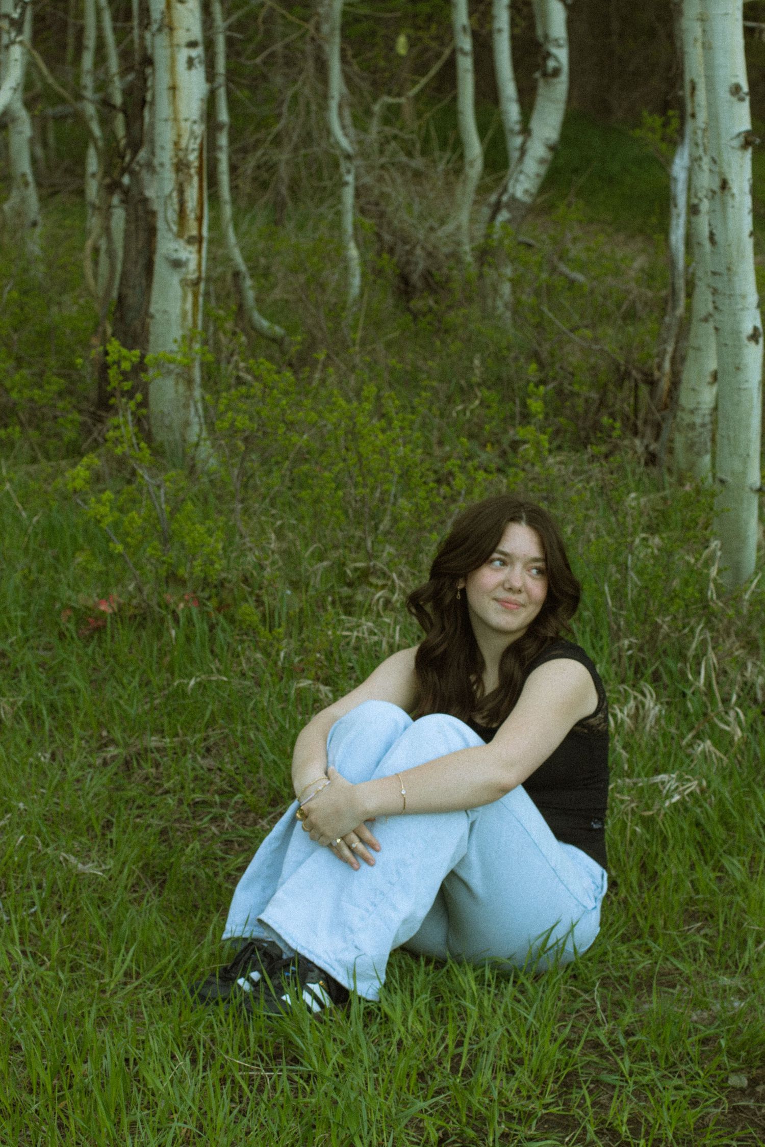 A person in a white top and jeans reclines on green grass in a wooded area with aspen trees in the background.