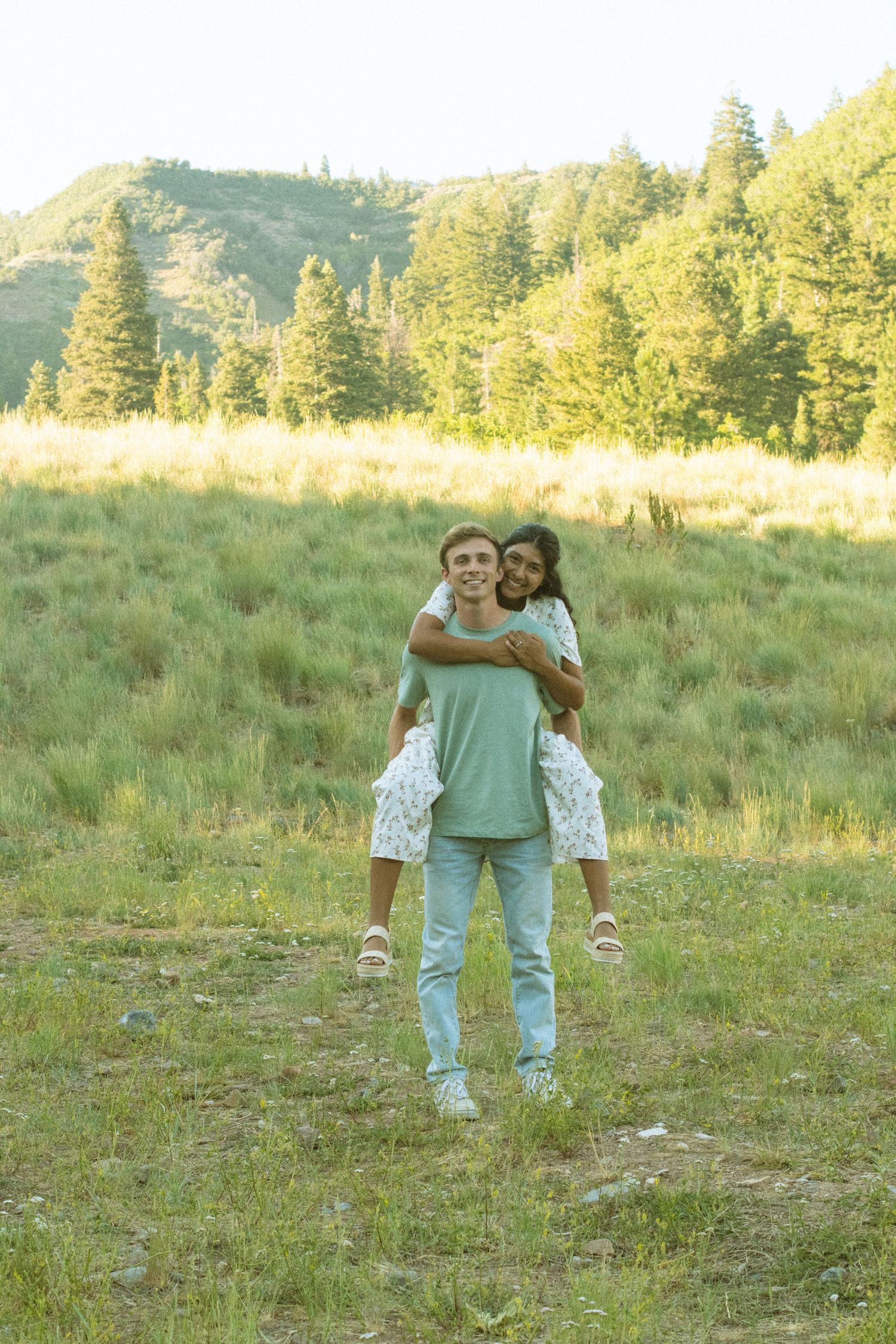 A couple enjoys a playful piggyback ride in a sunny mountain meadow surrounded by evergreen trees.