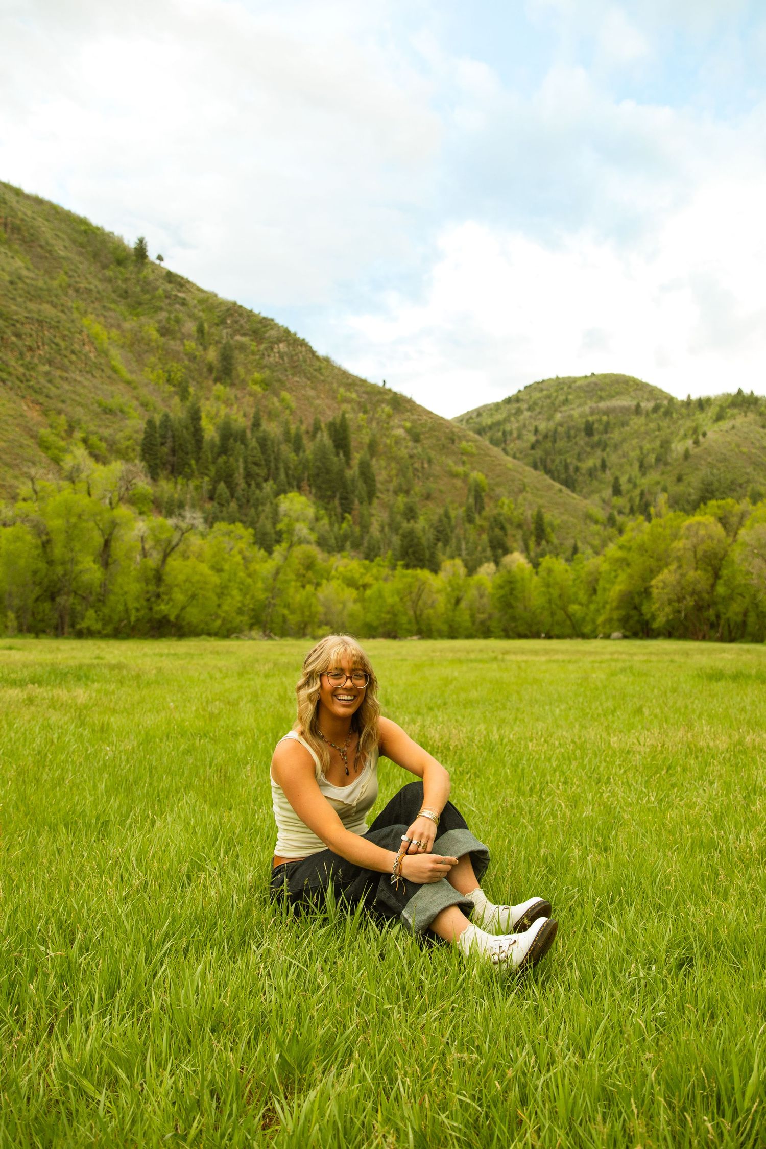 A smiling blonde woman sits cross-legged in a lush green meadow with tree-covered mountains under a cloudy sky.