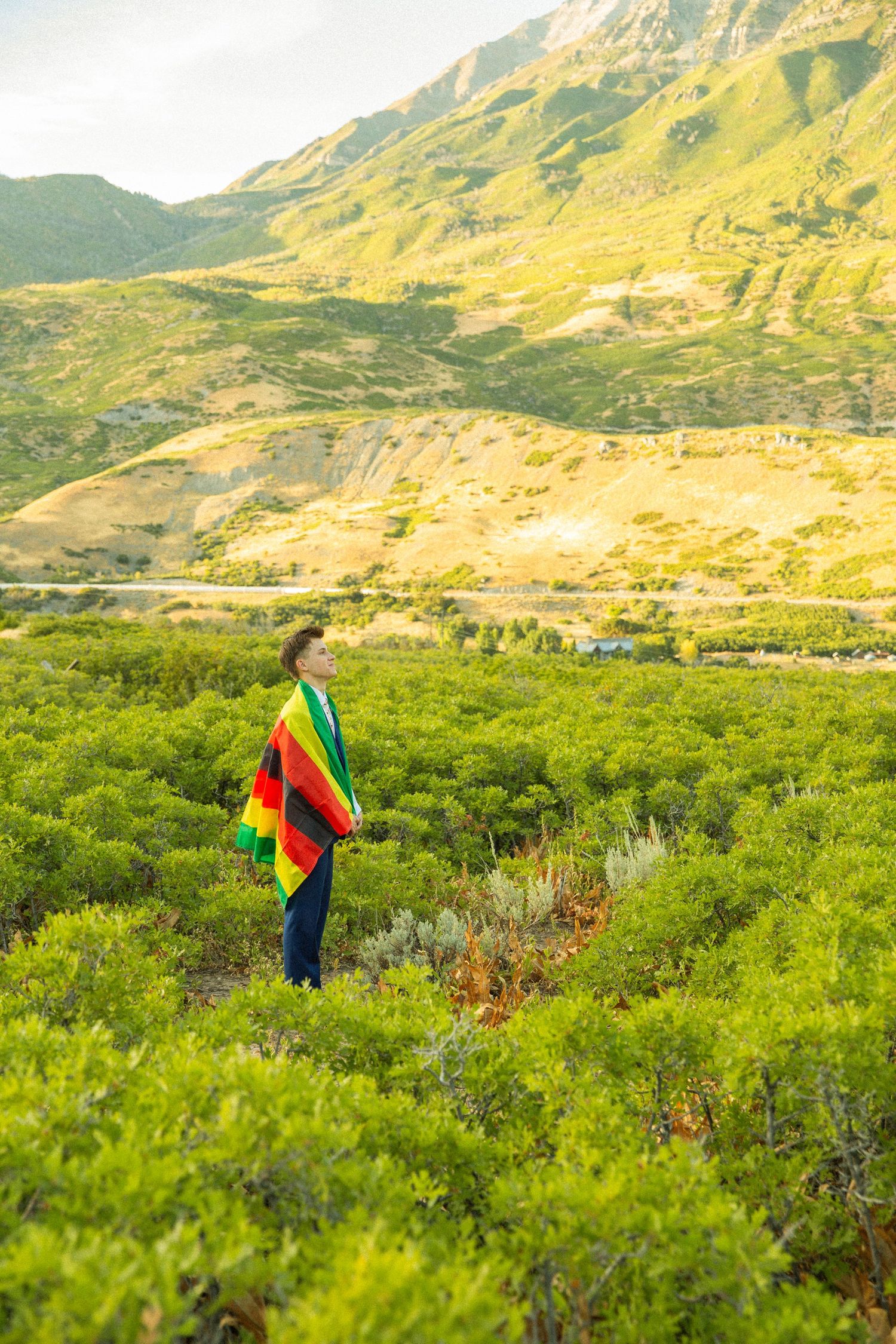 A figure wrapped in a colorful blanket stands in a green meadow with majestic mountains rising in the background.