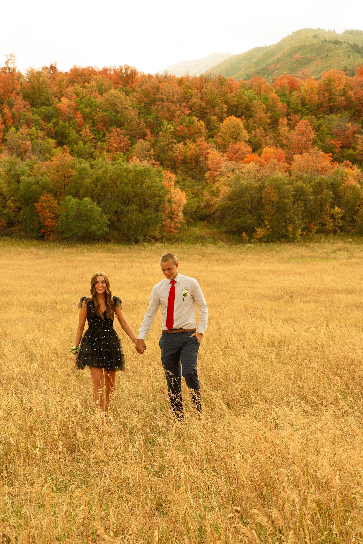 A couple walks hand in hand through a golden autumn meadow with vibrant orange foliage in the background.