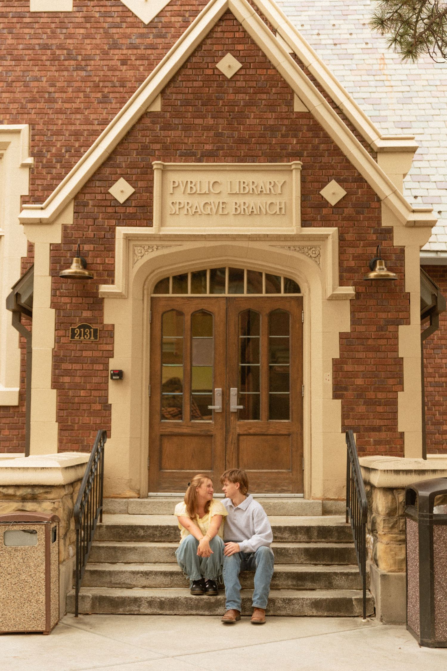 A couple sits on the steps of a historic brick public library building with cream-colored architectural details and arched doorway.