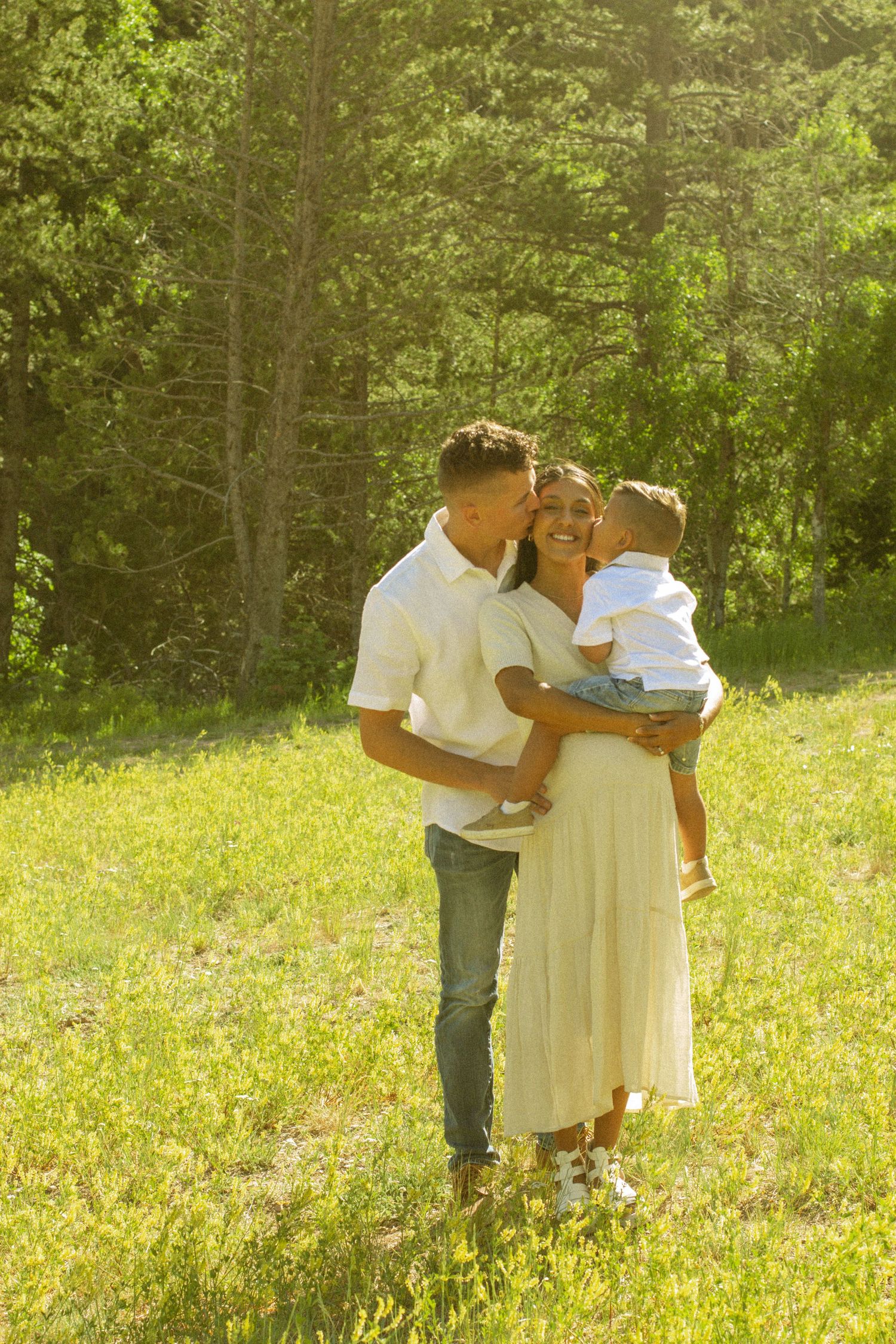 A couple shares a tender moment in a sunny meadow with lush green trees in the background.