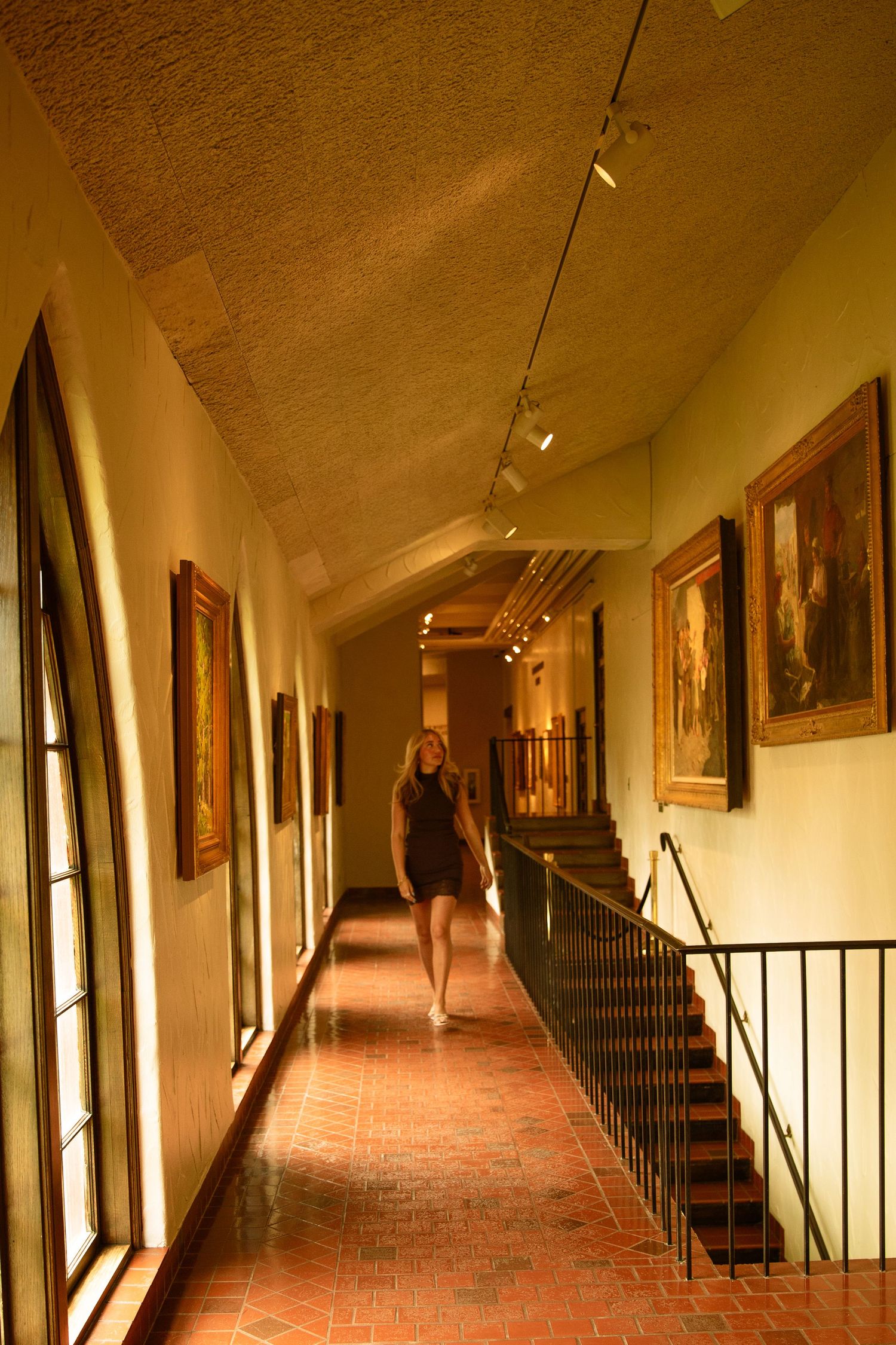 Woman walking through an elegant art gallery hallway with vaulted ceiling, track lighting, and paintings on warm golden walls.