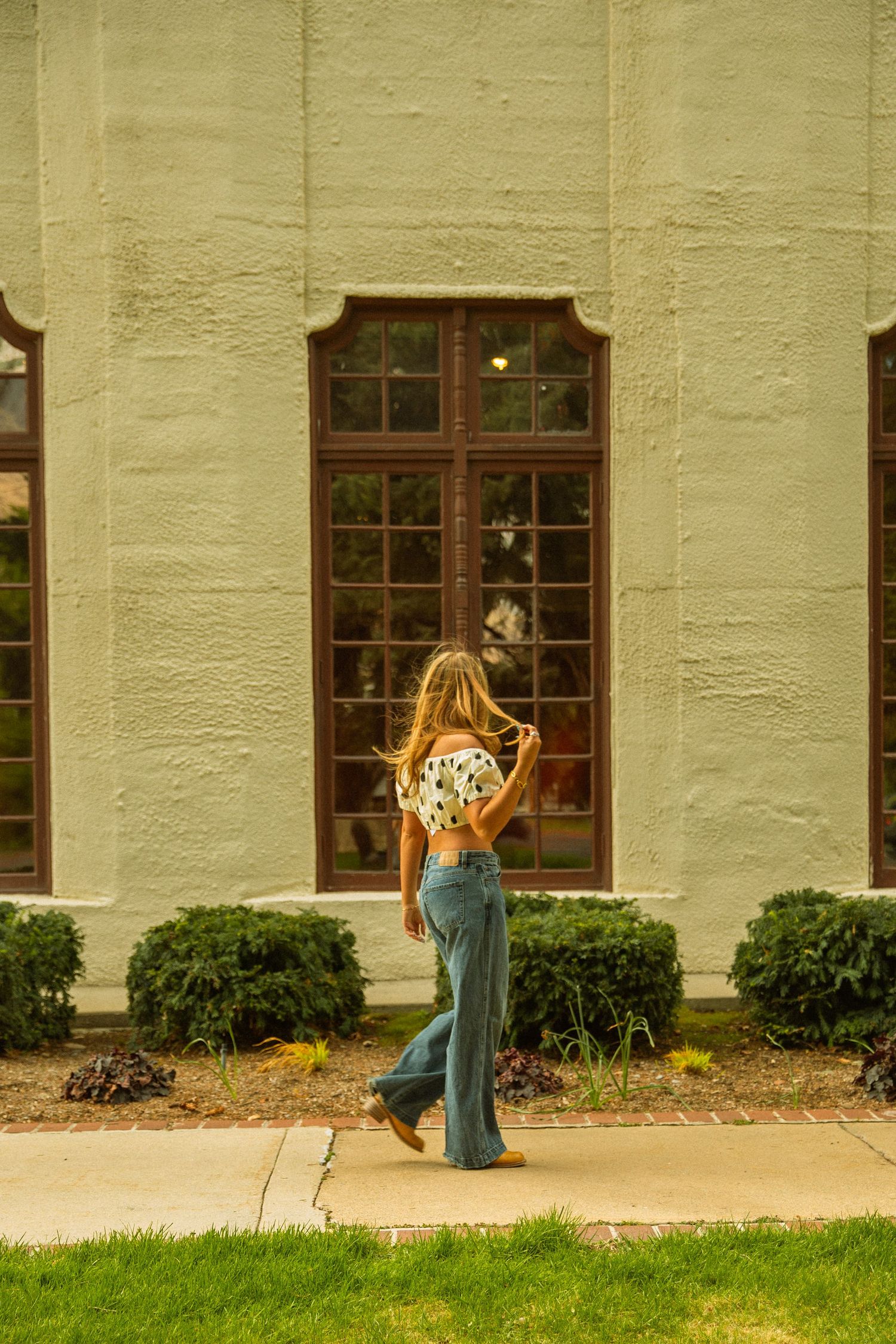 A woman in wide-leg jeans and crop top walks toward a green stucco building with arched windows.