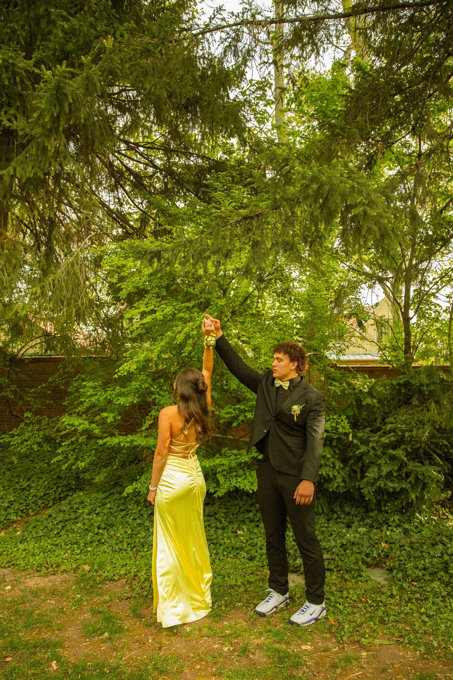 A couple in prom attire, yellow gown and dark suit, dancing outdoors among lush green trees.