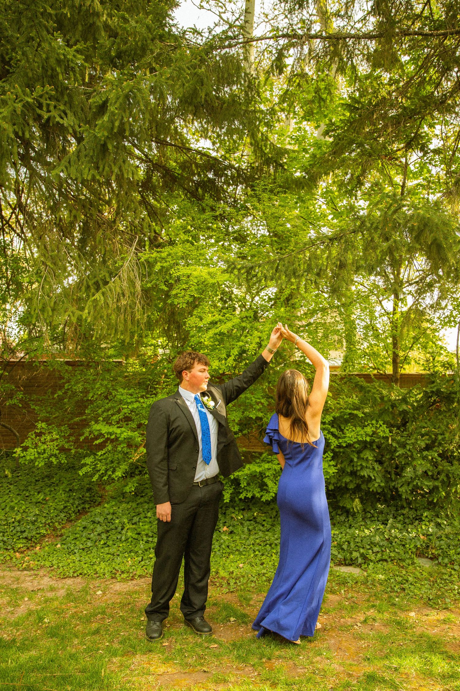 A couple in formal attire dancing outdoors, man in suit with blue tie and woman in elegant blue gown, surrounded by lush green trees.