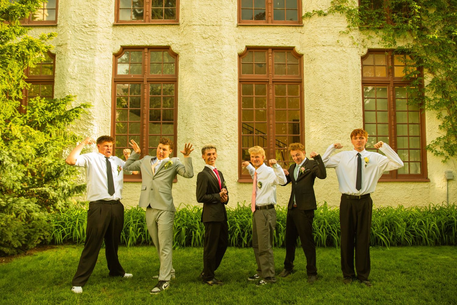 Six young men in formal attire flexing their muscles outdoors in front of a white brick building with large windows.
