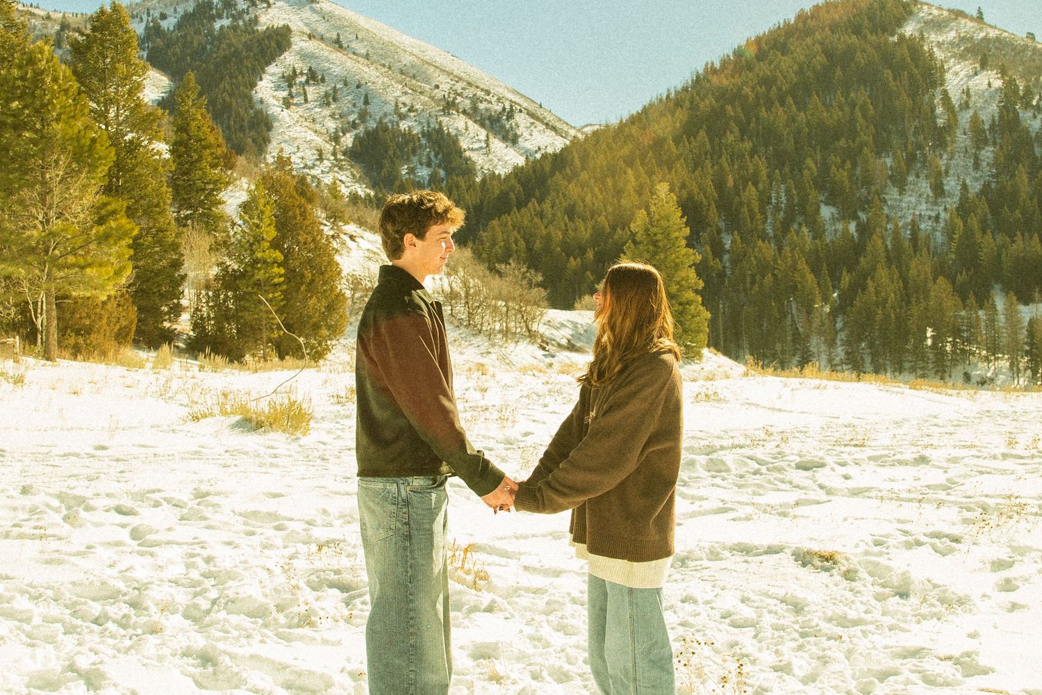 A couple shares a romantic moment in a snowy winter mountain landscape during sunset.