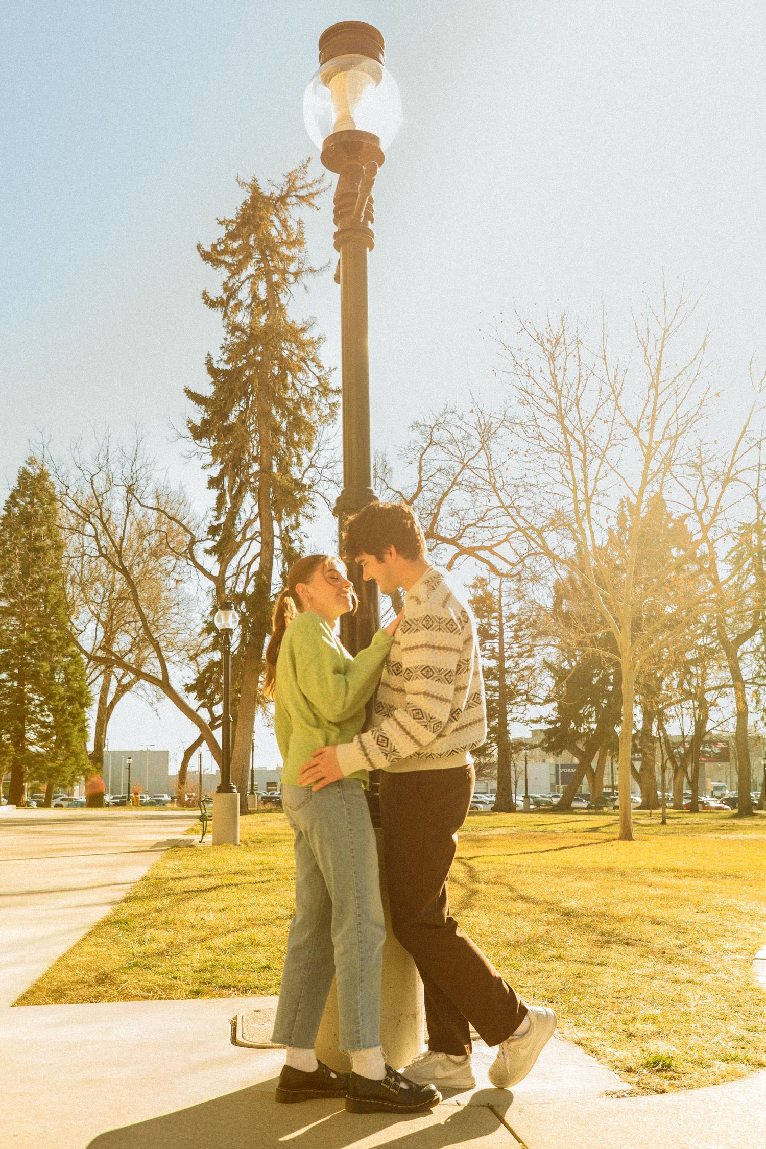 A couple embraces romantically under a vintage-style lamp post in a sunlit park on a winter afternoon.
