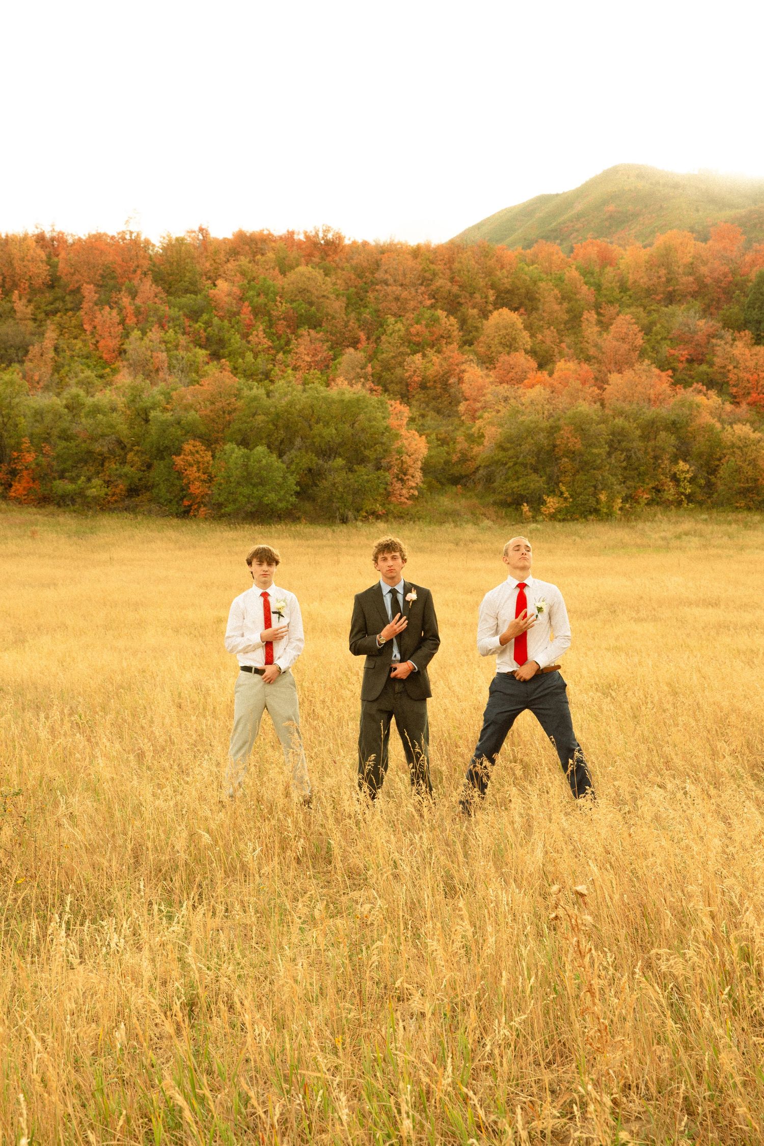 Three groomsmen in suits and red ties walk through a golden autumn field with fall foliage in the background.