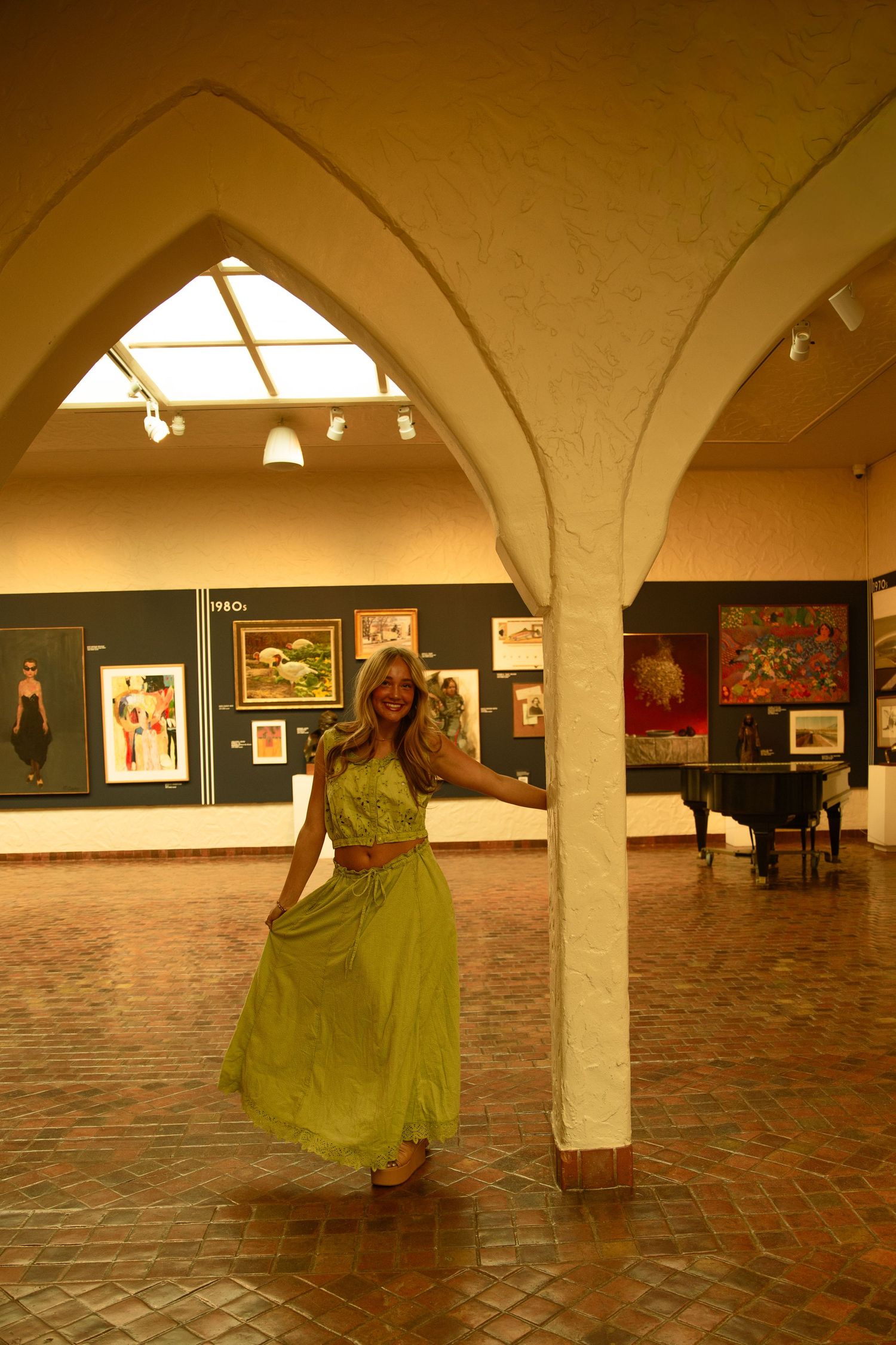 A woman in a green dress poses gracefully in an art gallery with arched columns and colorful paintings on display.
