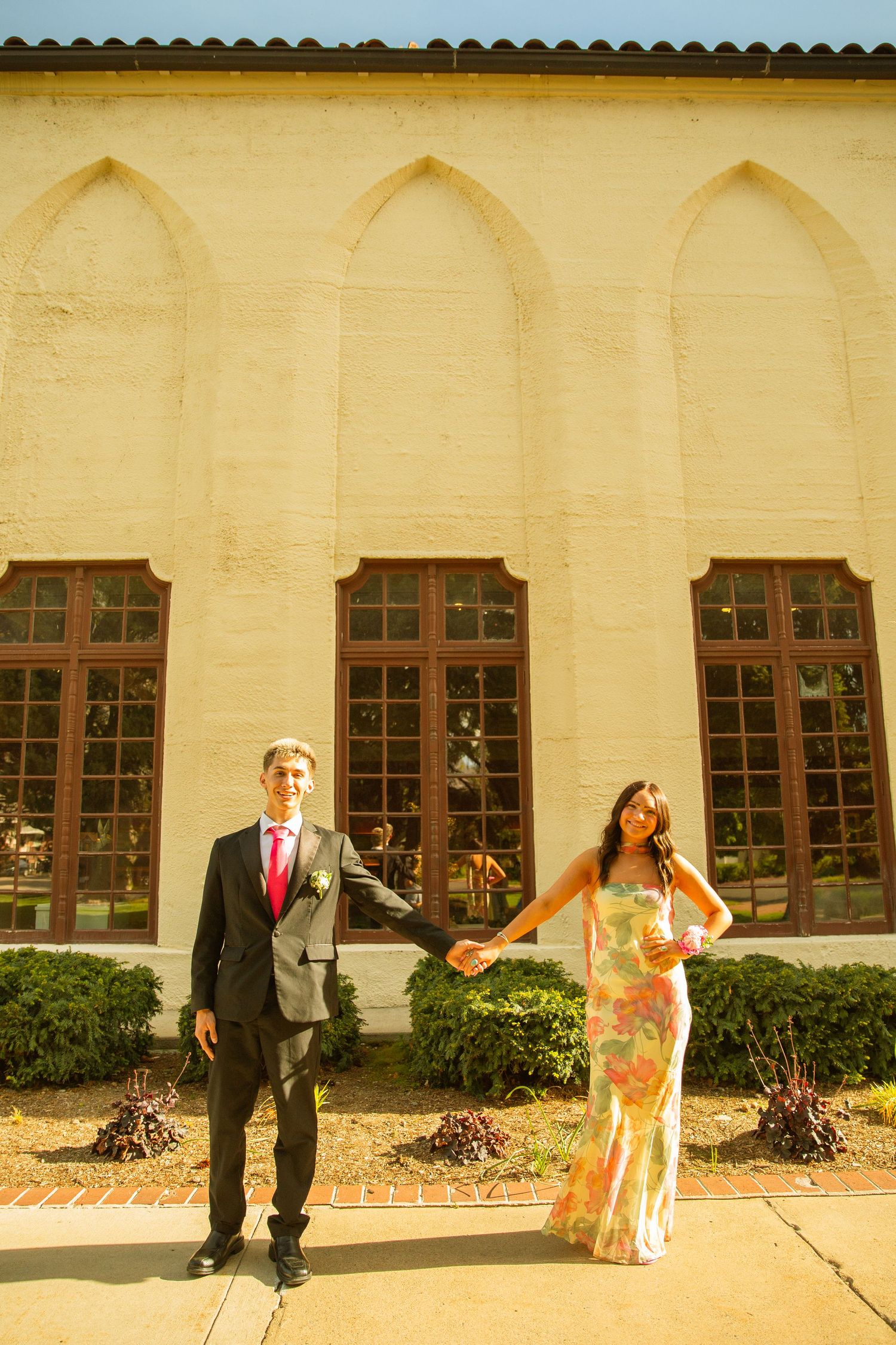 A couple in formal attire holds hands outside a historic building, the man in a suit and woman in a floral gown.