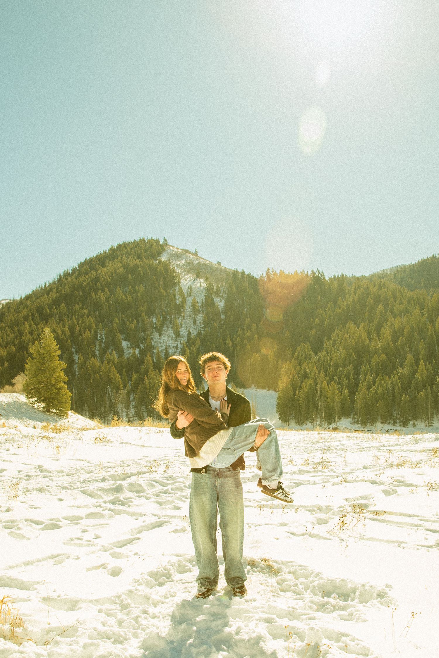 A person plays in deep snow on a mountain slope with evergreen trees and bright sunlight in the background.