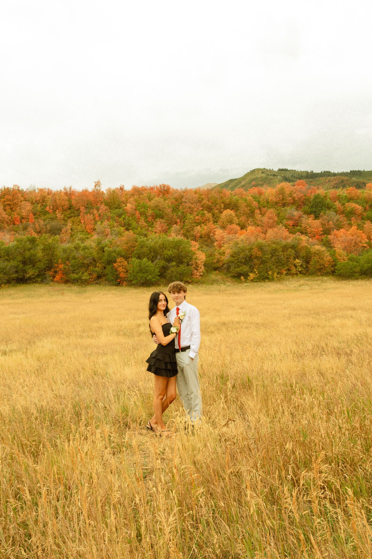 A couple shares an intimate moment in a golden wheat field during autumn with colorful trees in the background.