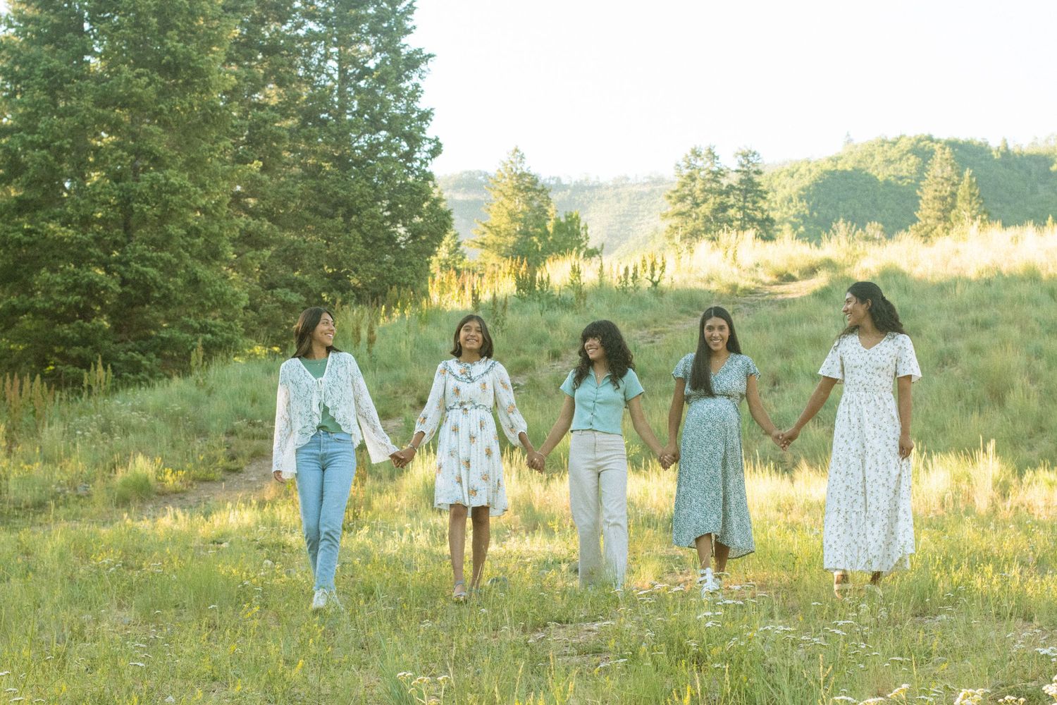 Five people in coordinating blue and white outfits hold hands while walking through a sunny meadow with evergreen trees.