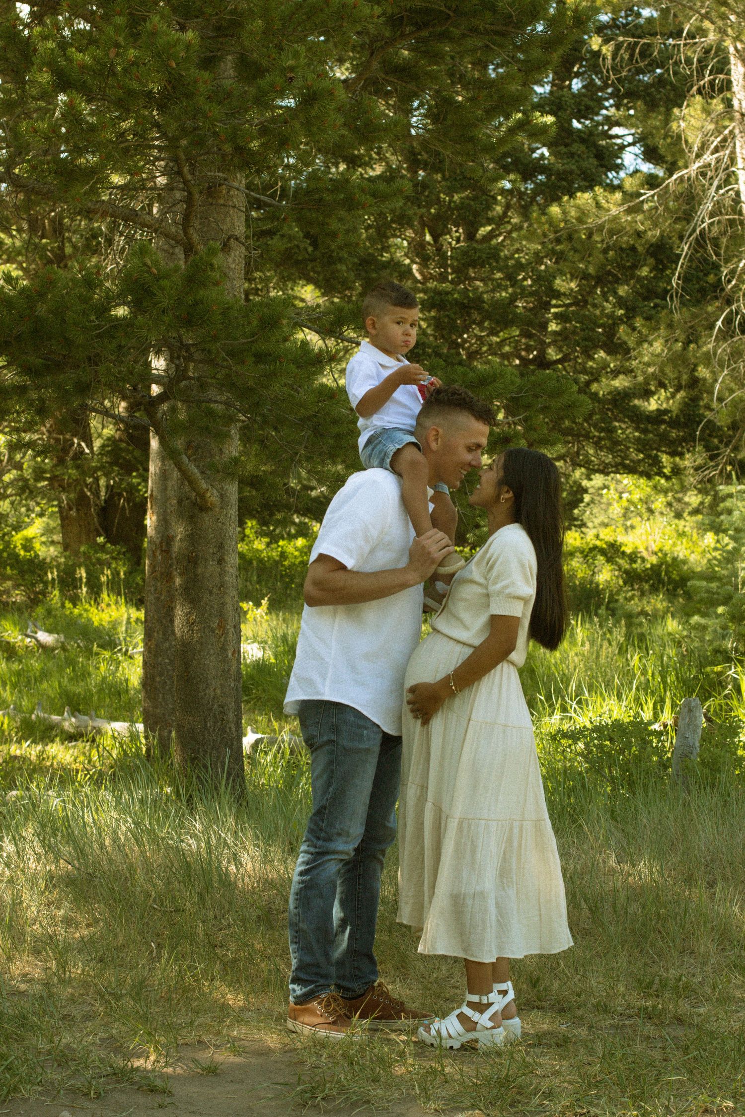 A couple shares a tender moment in a sunlit forest setting while wearing casual white and denim outfits.