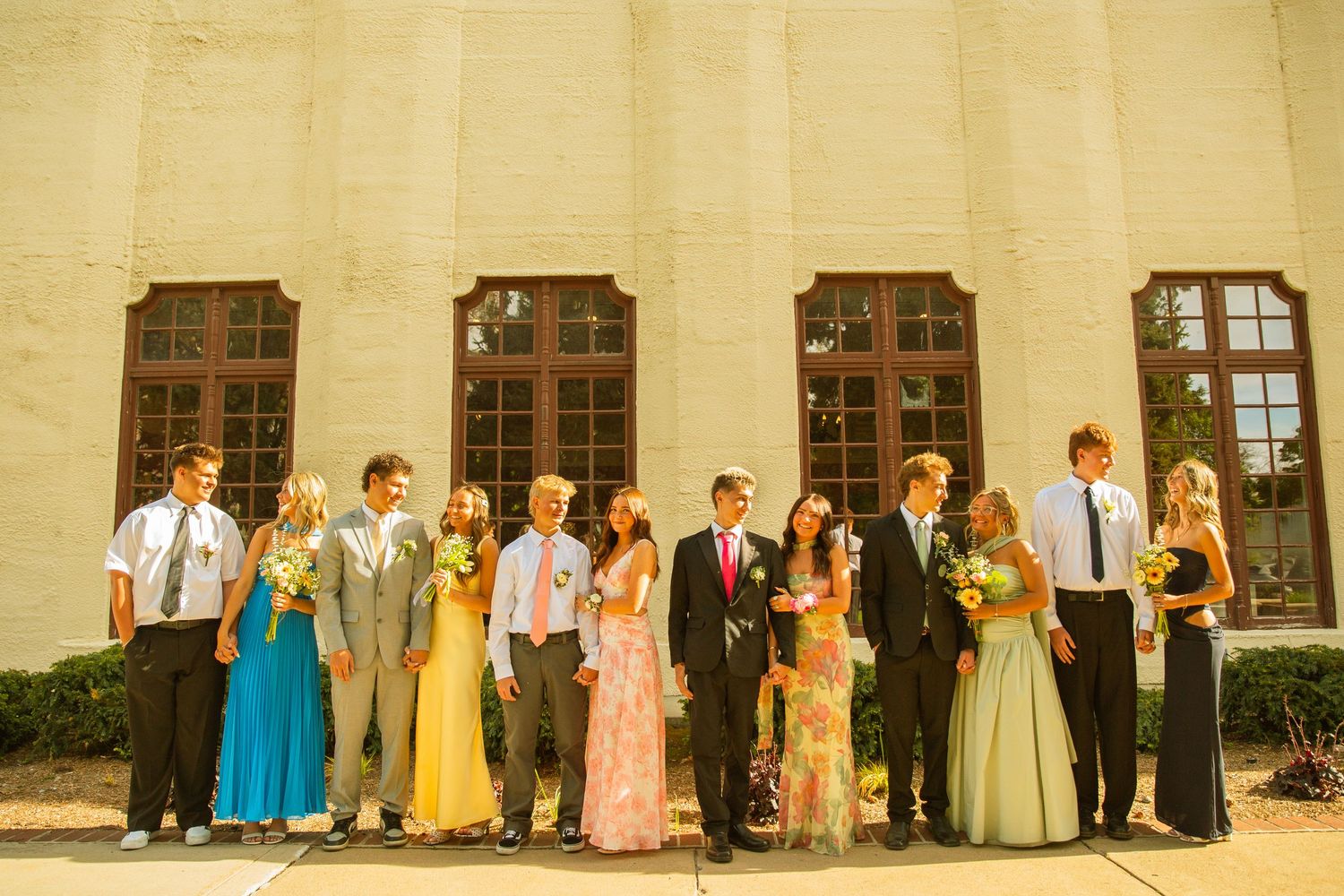 A group of prom couples posing in formal attire outside a brick building with large windows.