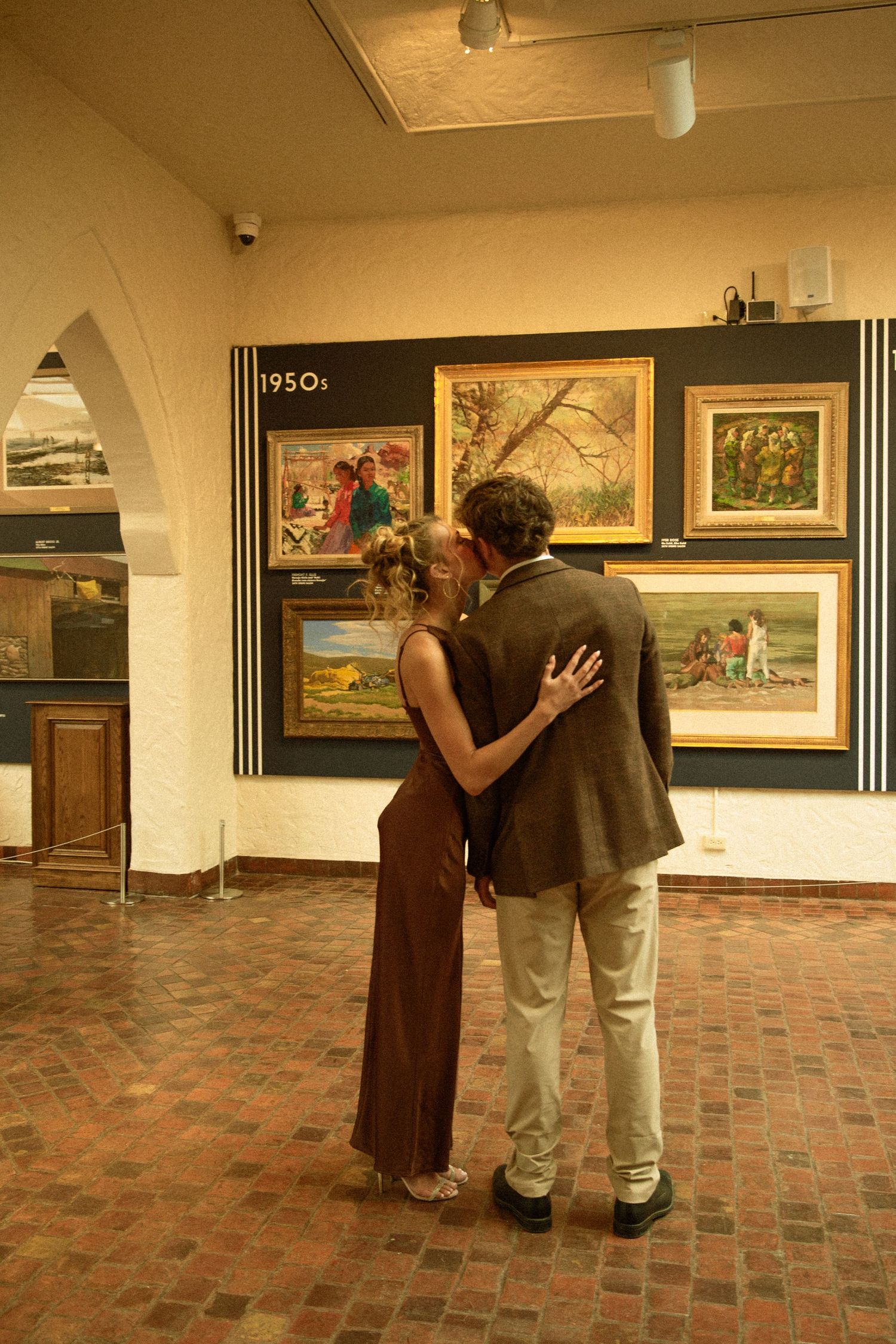 Two people examine artwork displayed on gallery walls while standing on a brick floor in an art exhibition space.