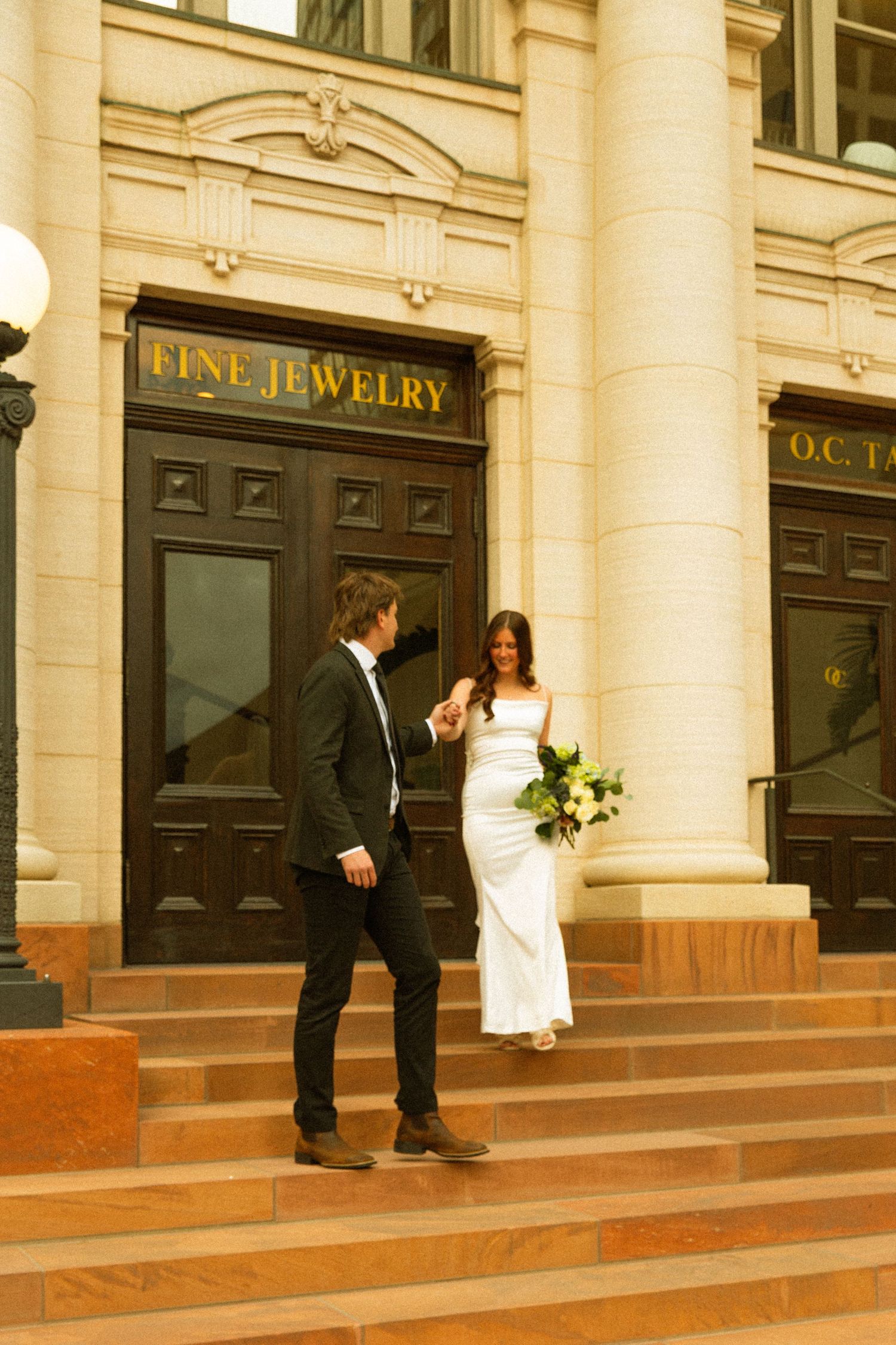 A couple in wedding attire stands on the steps of a historic building with a Fine Jewelry storefront.
